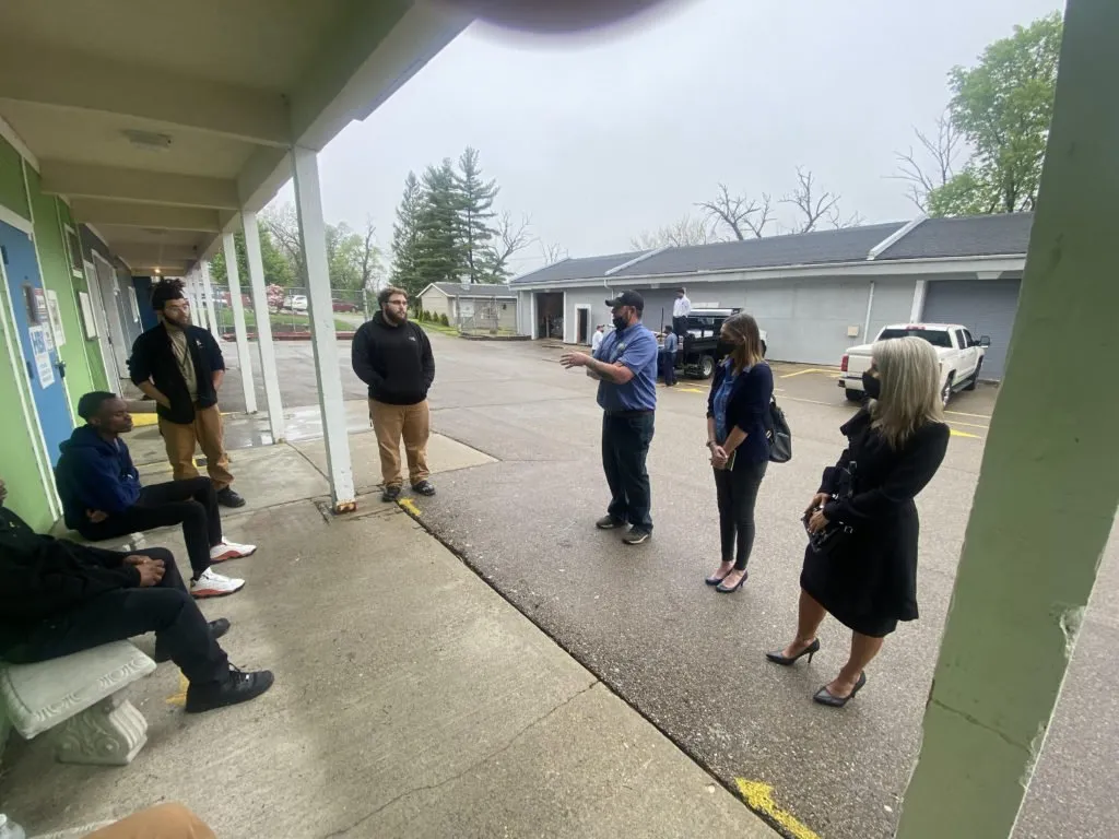 A group of individuals standing and sitting outside a building, engaged in a discussion. The scene captures a mix of people, including those in casual clothing and a woman in a black dress, all under a covered walkway.
