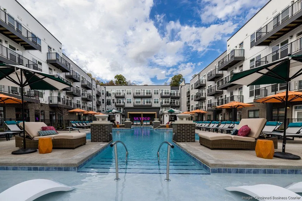 A vibrant pool area surrounded by modern apartment buildings, featuring lounge chairs and orange umbrellas under a bright sky.