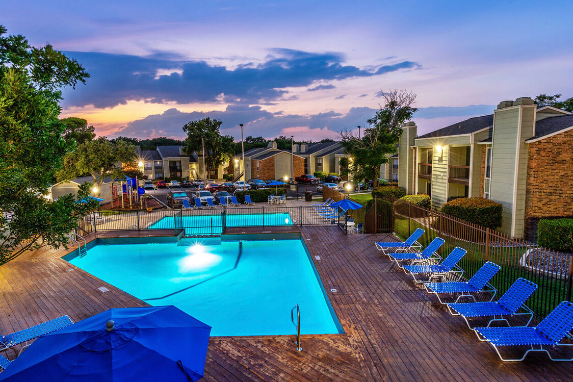 A view of a well-lit swimming pool area surrounded by lounge chairs and umbrellas at dusk.