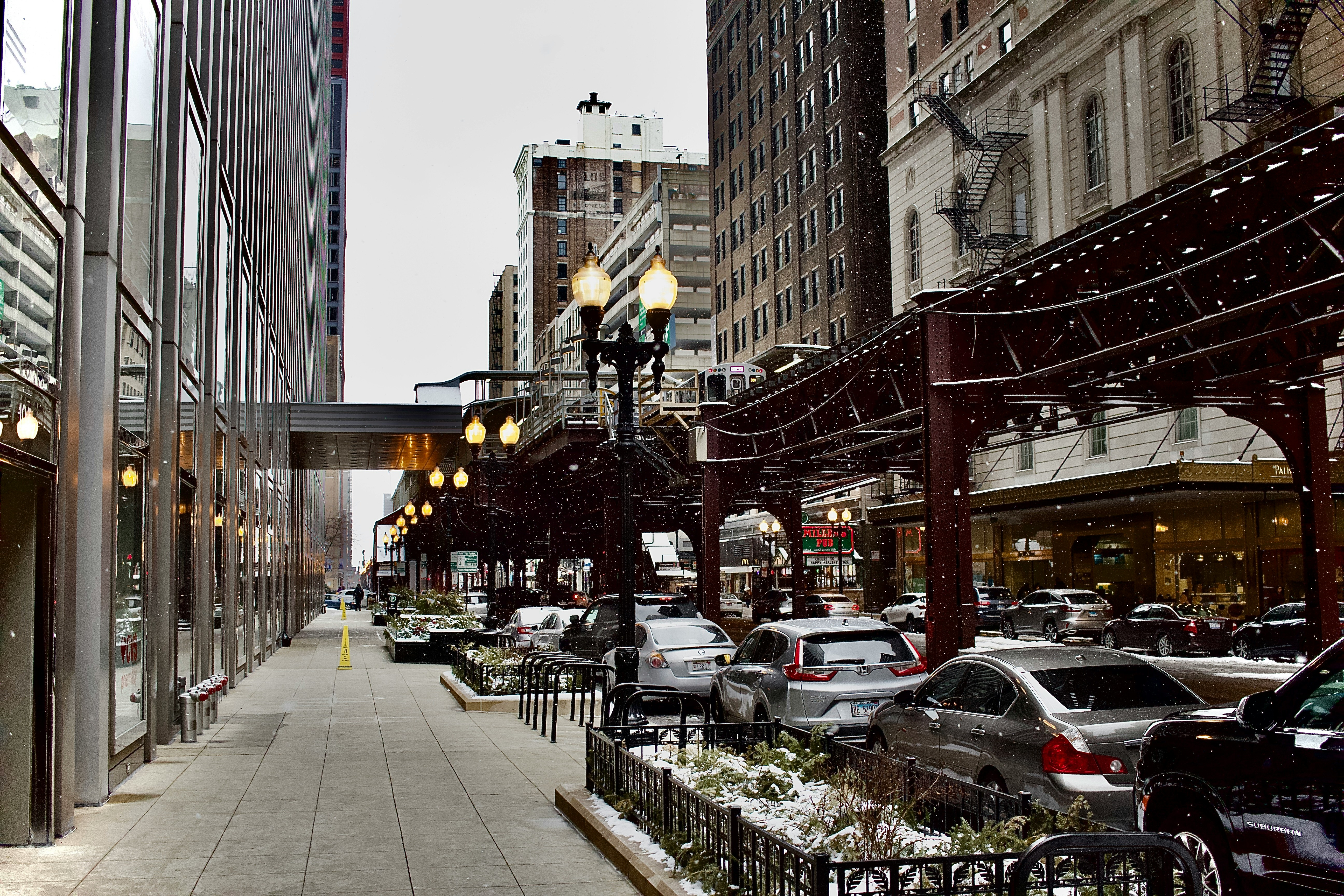 A snowy city street with vehicles parked along the curb, surrounded by tall buildings.