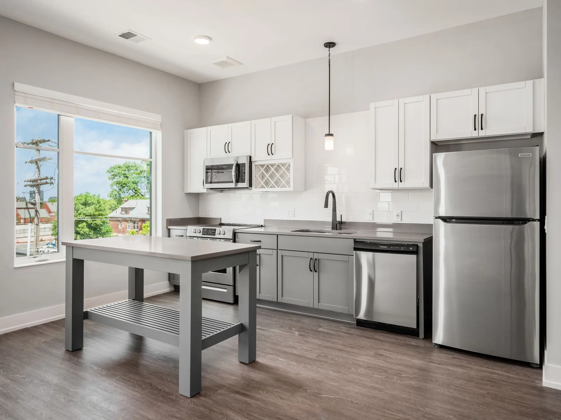 A modern kitchen featuring gray cabinetry, stainless steel appliances, and a center table with a wooden surface and gray legs. Large window providing natural light.