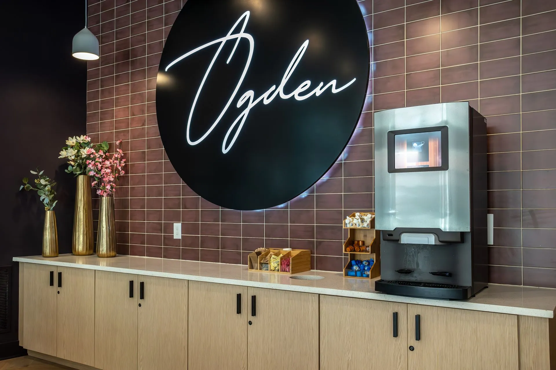 A modern communal area featuring a large circular sign that says 'Ogden', a beverage dispenser, and decorative plants in stylish vases.