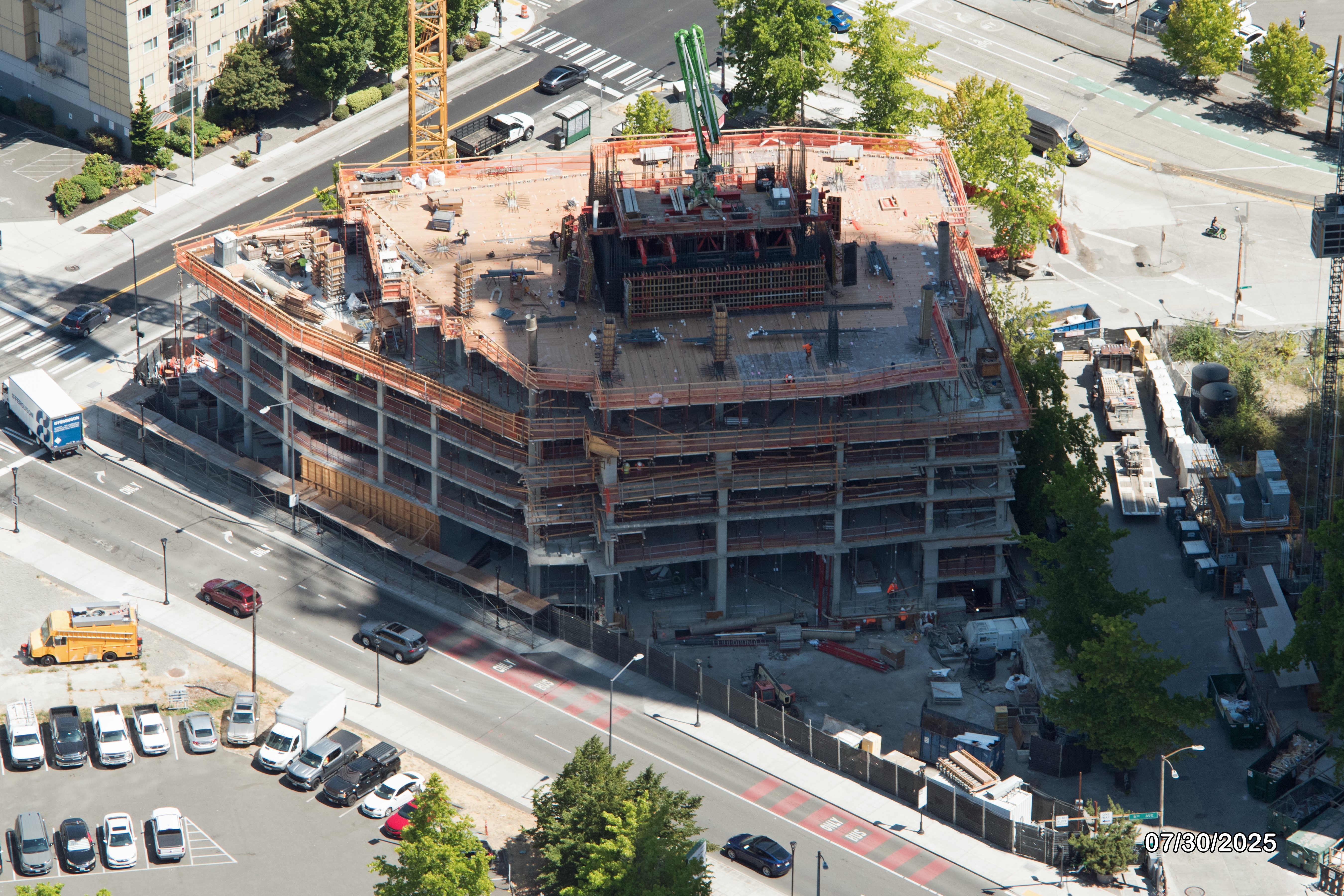 Aerial view of an under-construction building with cranes, surrounded by trees and parked cars on the street.