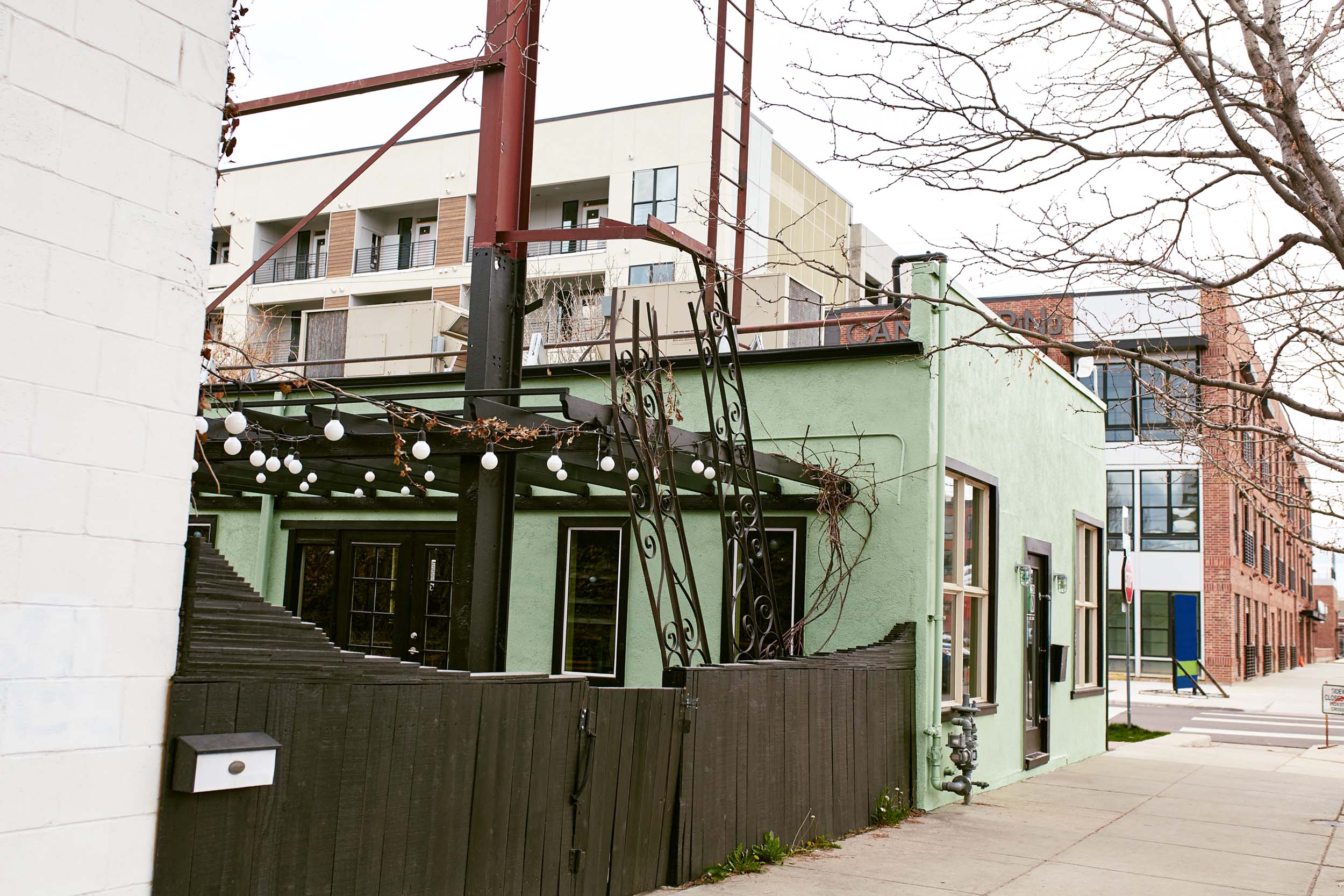 A street view showing a green building with outdoor string lights and a wooden fence, adjacent to a modern multi-story apartment building in the background.