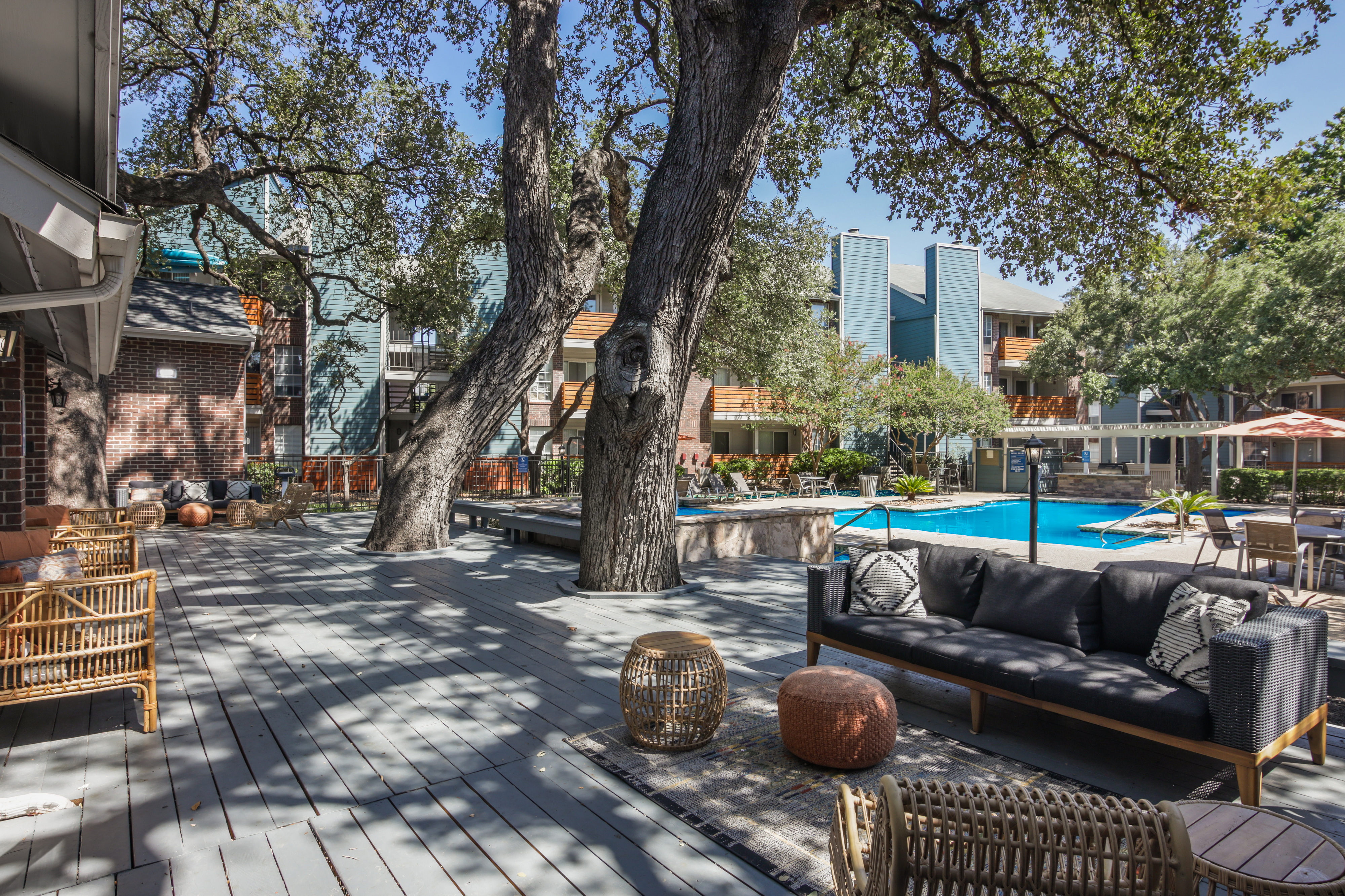 A spacious outdoor living area featuring shaded seating, large trees, and a swimming pool in the background.
