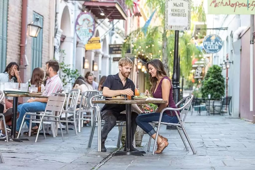 A couple enjoying a meal at an outdoor café on a charming street, surrounded by greenery and other diners.