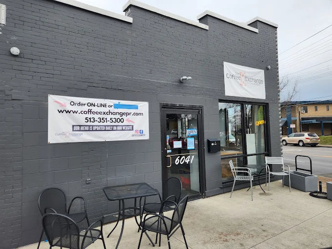Exterior of Coffee Exchange, a coffee shop with a gray wall and a sign. There are tables and chairs outside and a banner promoting online orders.