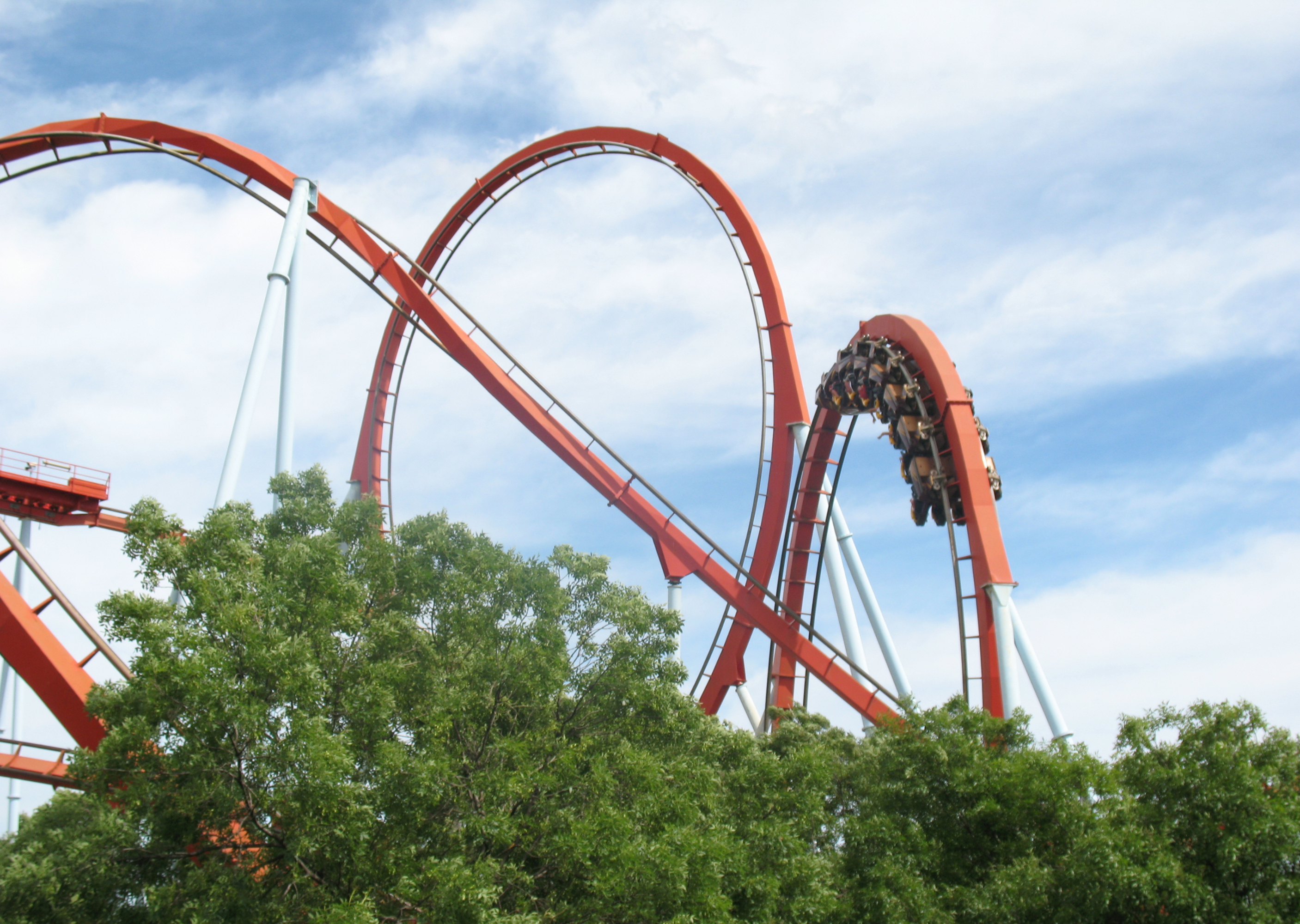 A roller coaster with bright red tracks looping through a blue sky, partially obscured by lush green trees.
