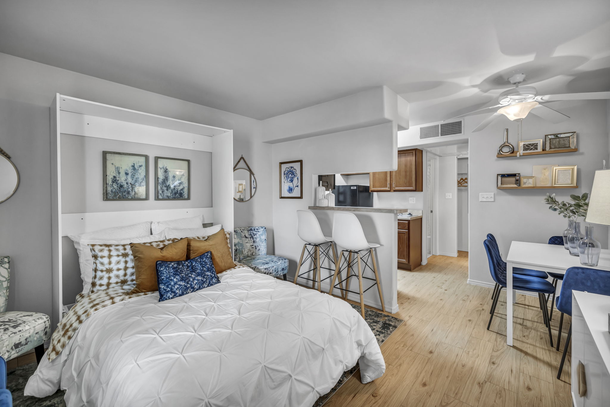 A stylish studio apartment featuring a bed with white bedding and decorative pillows, a small dining area with blue chairs, a kitchenette, and modern decor.