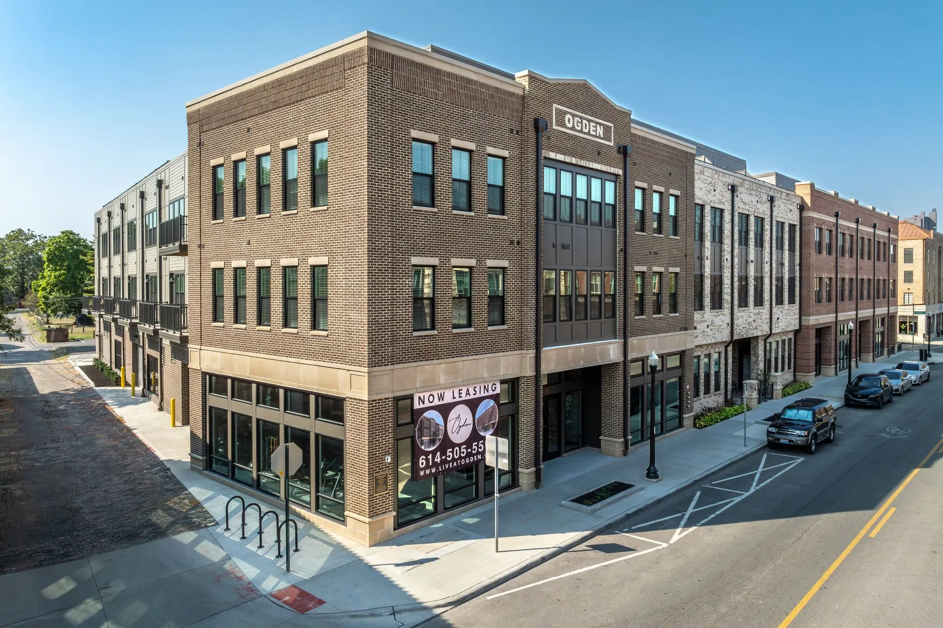 Exterior view of a multi-story commercial building with brick facade and large windows, featuring a 'Now Leasing' sign at the entrance.