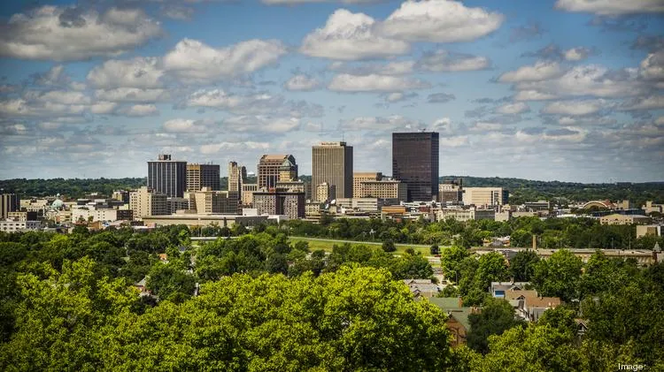 A panoramic view of a city skyline featuring tall buildings under a partly cloudy sky, surrounded by greenery.