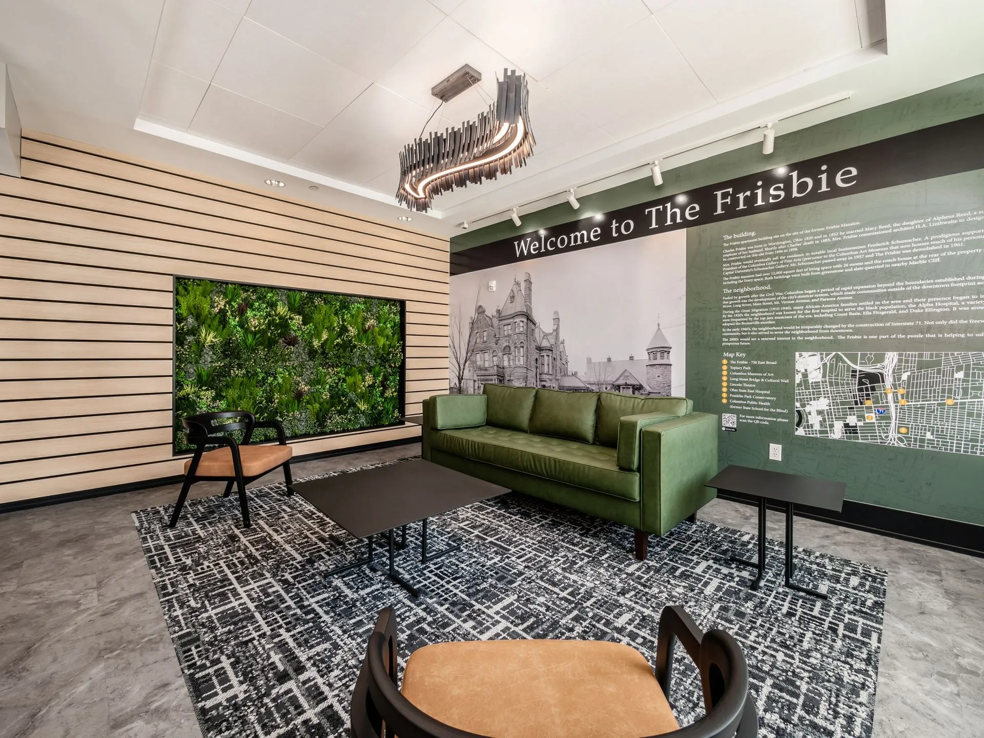 A modern community lounge with a green couch, two chairs, and decorative elements including a vertical garden and a large black and white mural.