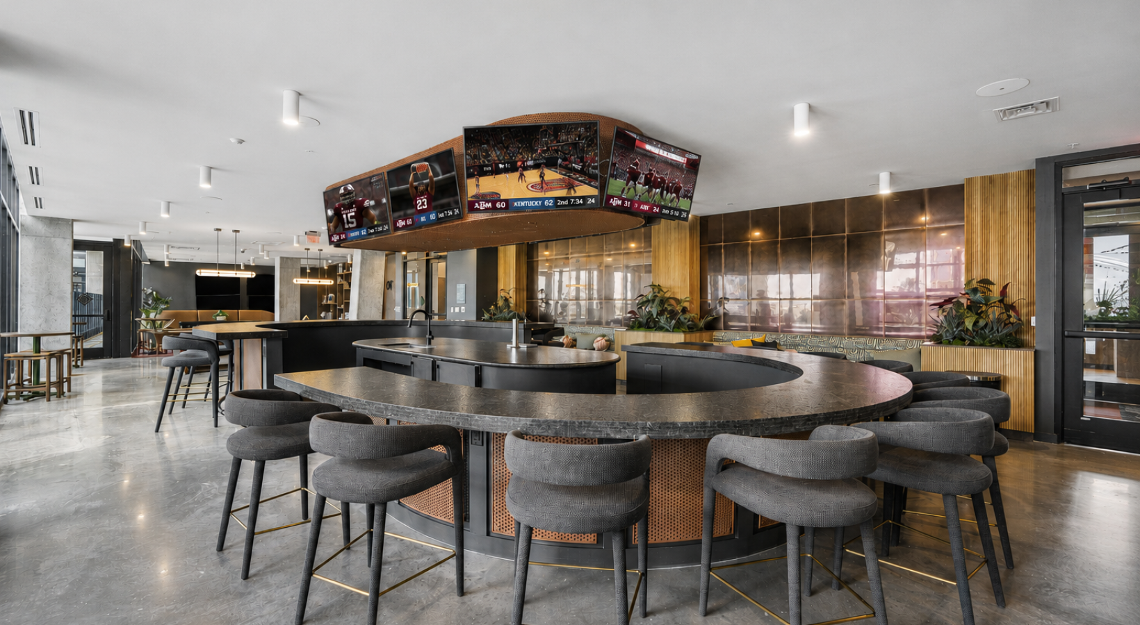 A modern community lounge featuring a curved bar with high chairs, multiple screens displaying sports events, and contemporary decor with plants.