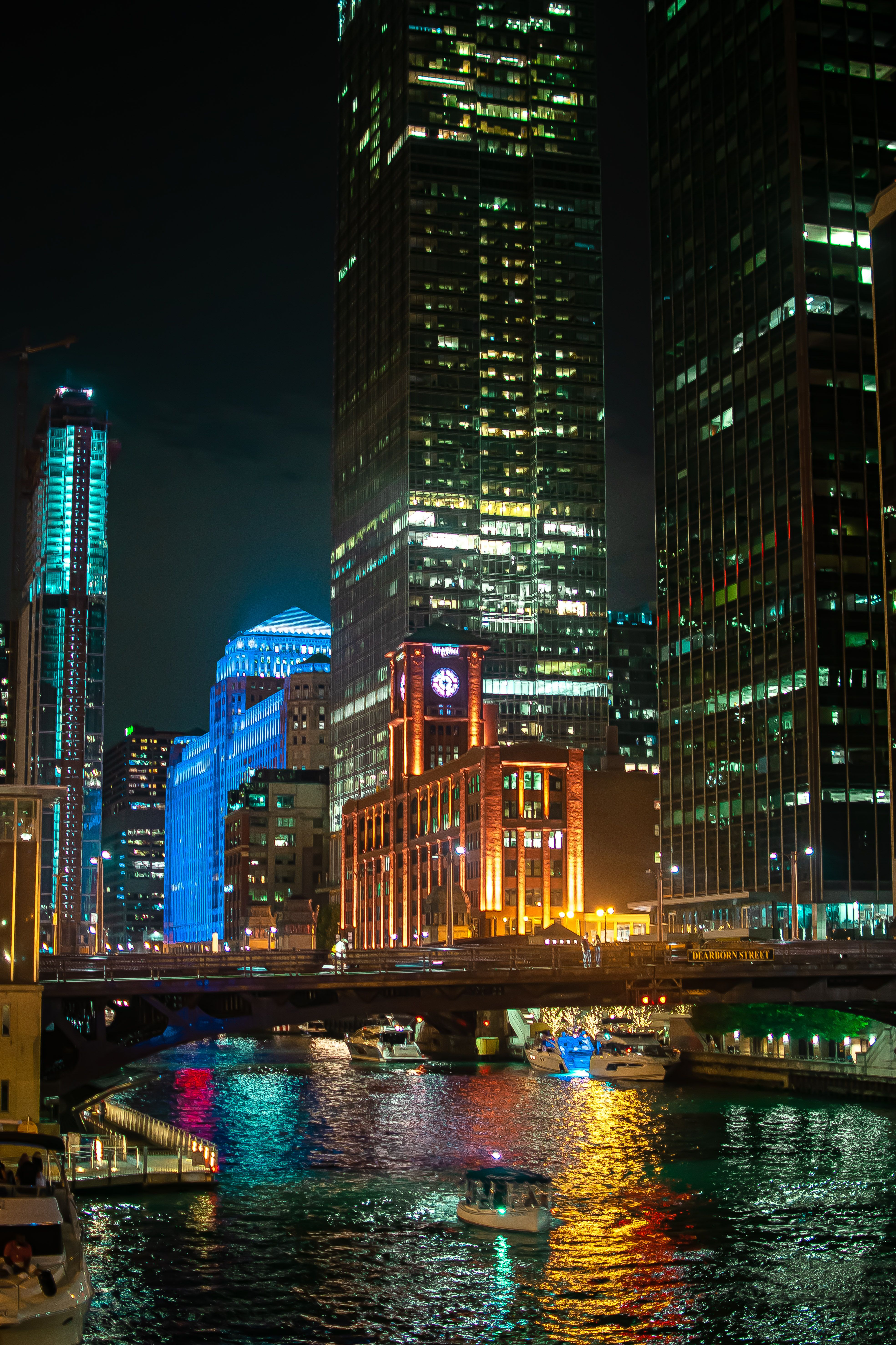 A vibrant city skyline at night, showcasing tall buildings illuminated with colorful lights next to a river filled with boats.