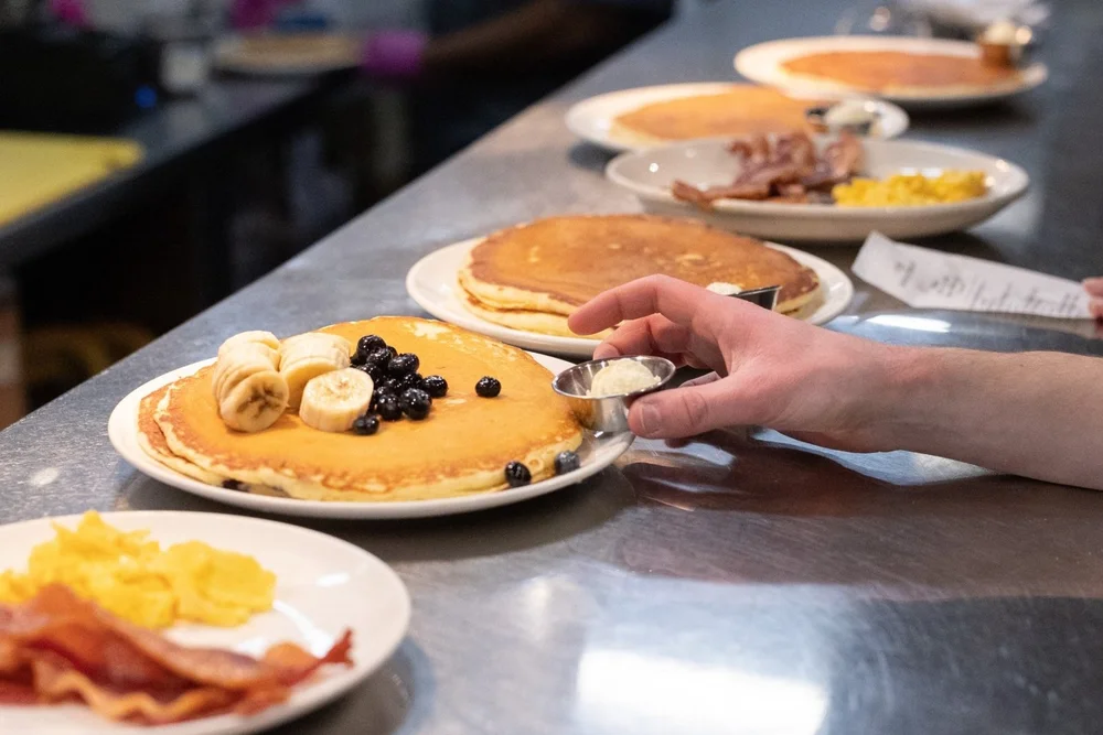 A hand about to sprinkle a topping on a stack of pancakes with bananas and blueberries, alongside other plates of pancakes and breakfast items arranged on a counter.