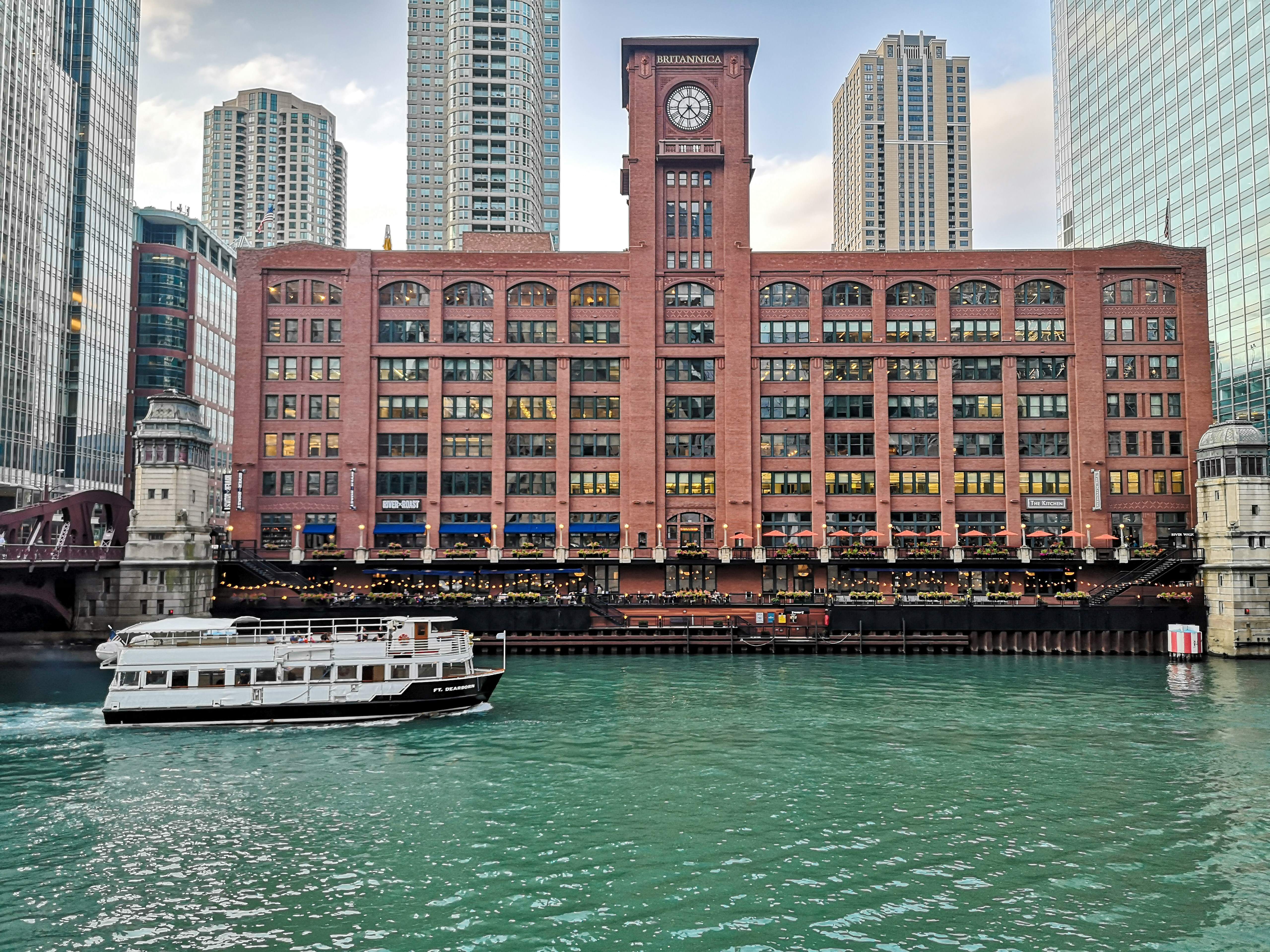 A historic red brick building with a clock tower along a river, surrounded by modern skyscrapers.