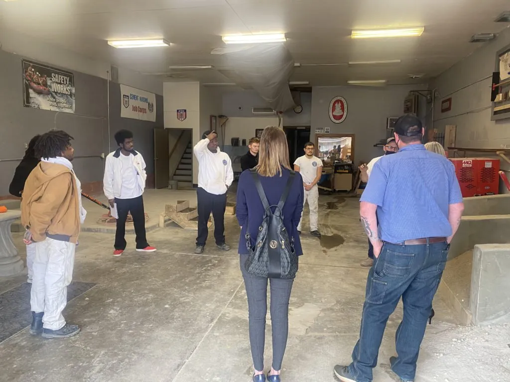 A group of people gathered in an indoor space discussing or participating in an activity. Some individuals are wearing white uniforms, while others are in casual attire. There are various tools and equipment visible in the background.