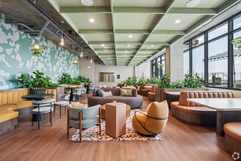 A stylish shared community lounge with a mix of seating options including colorful couches, chairs, and tables surrounded by greenery and large windows.