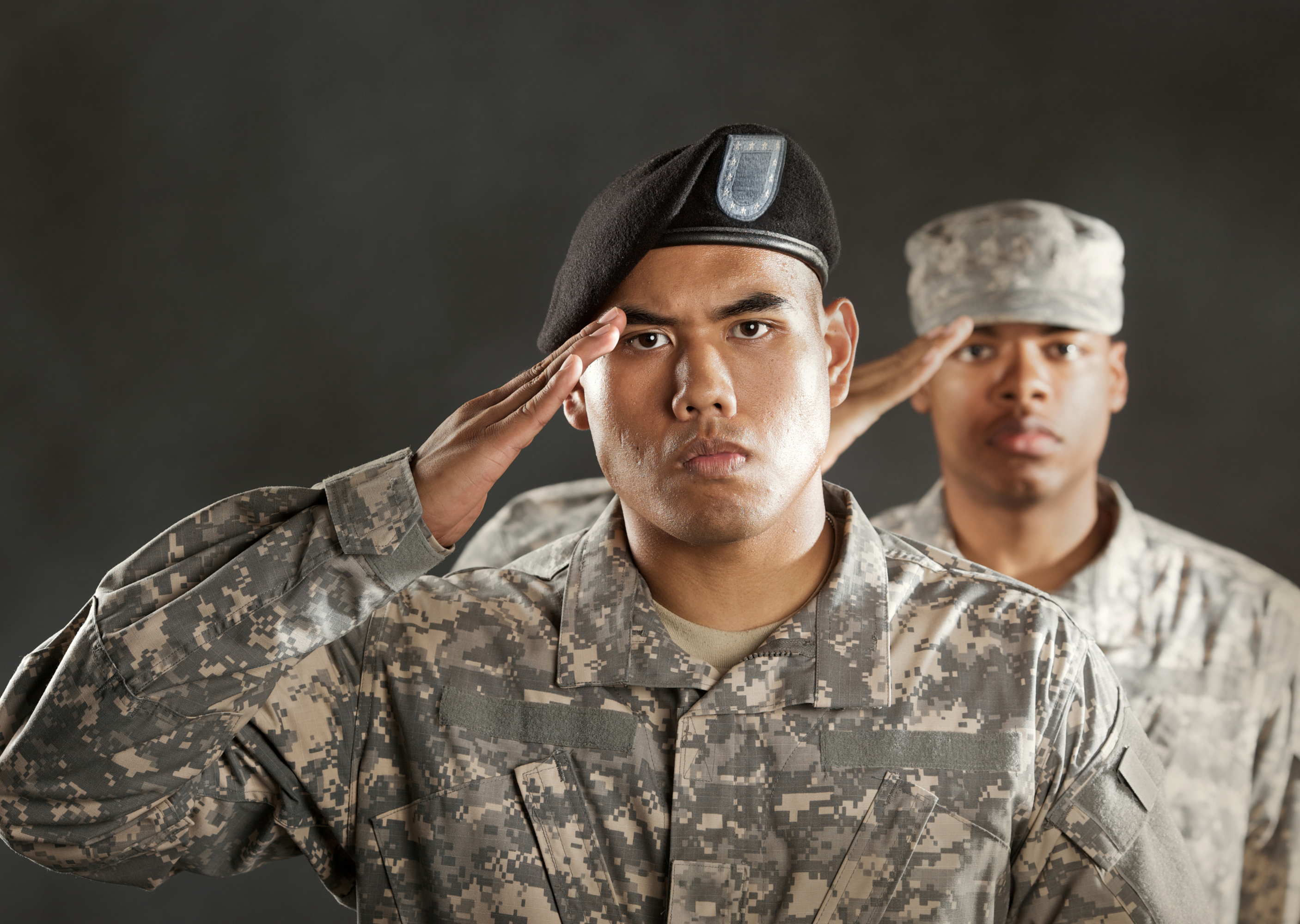 Two soldiers in military uniforms saluting, one in front wearing a beret and the other wearing a camouflage cap.