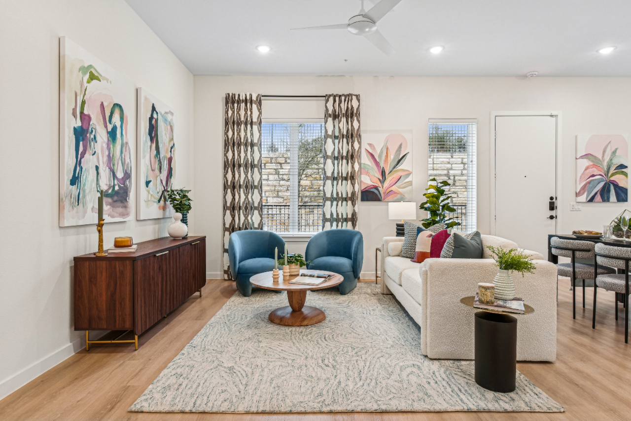 A stylish living room featuring two blue accent chairs, a white sofa, and a round coffee table on a light rug. The walls are adorned with colorful artwork, and there are large windows with patterned curtains. A dark wood side table and potted plants add to the decor.