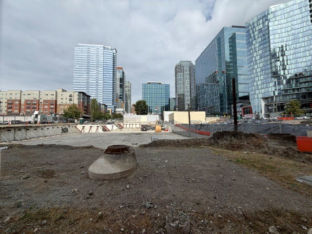 An urban construction site with gravel ground, surrounded by modern tall buildings under a cloudy sky.