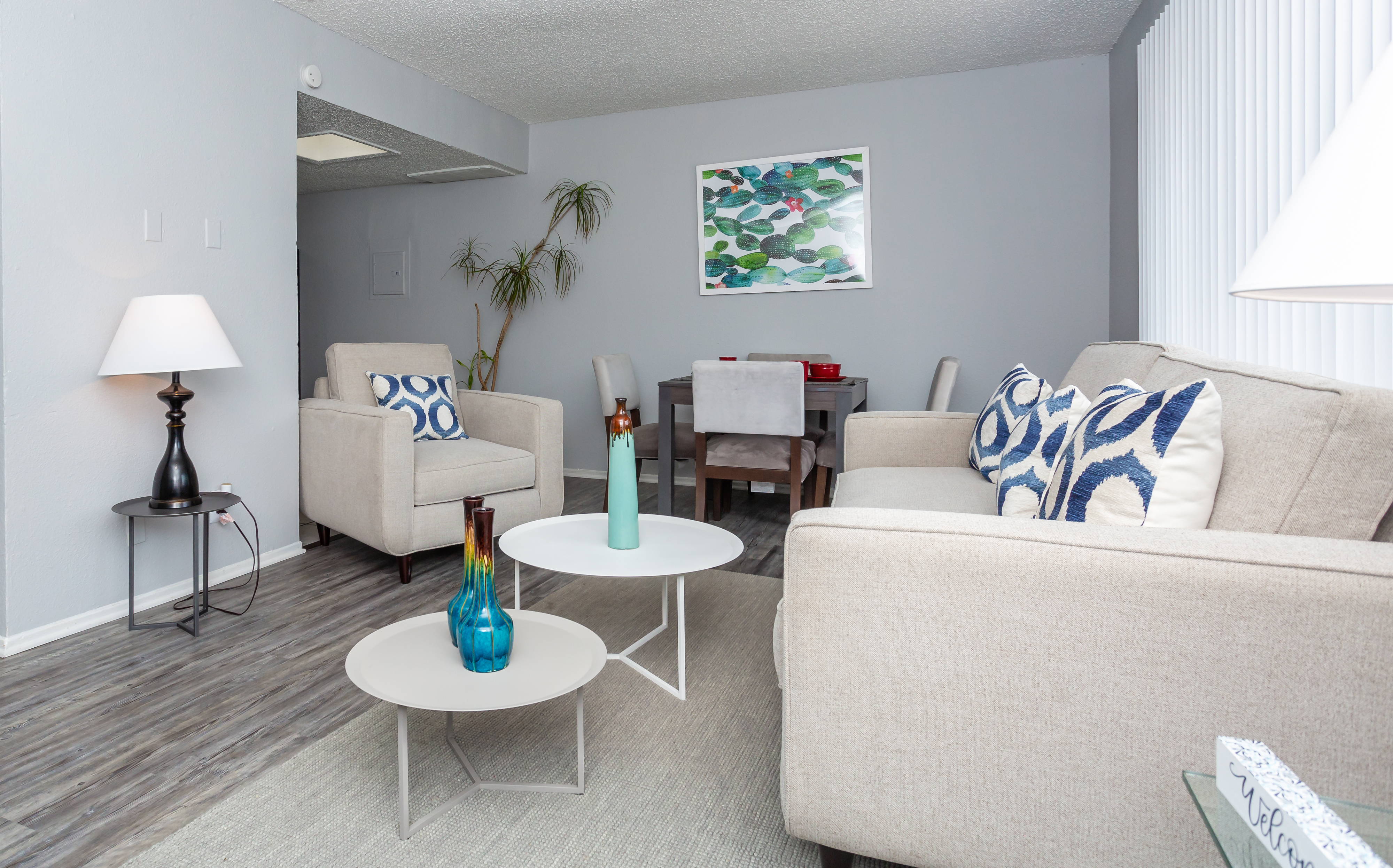 A modern living room featuring beige sofas with blue and white patterned cushions, a round white coffee table, and colorful decorative vases. There's a small dining area in the background with a dark wood table and chairs, and a large window with vertical blinds allowing natural light in.