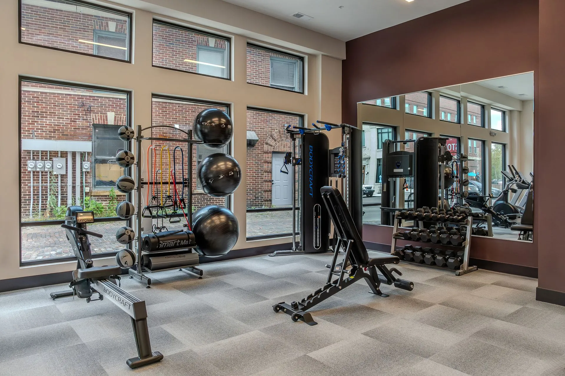 A modern apartment gym featuring various fitness equipment including weights, a weight machine, and exercise balls near large windows.