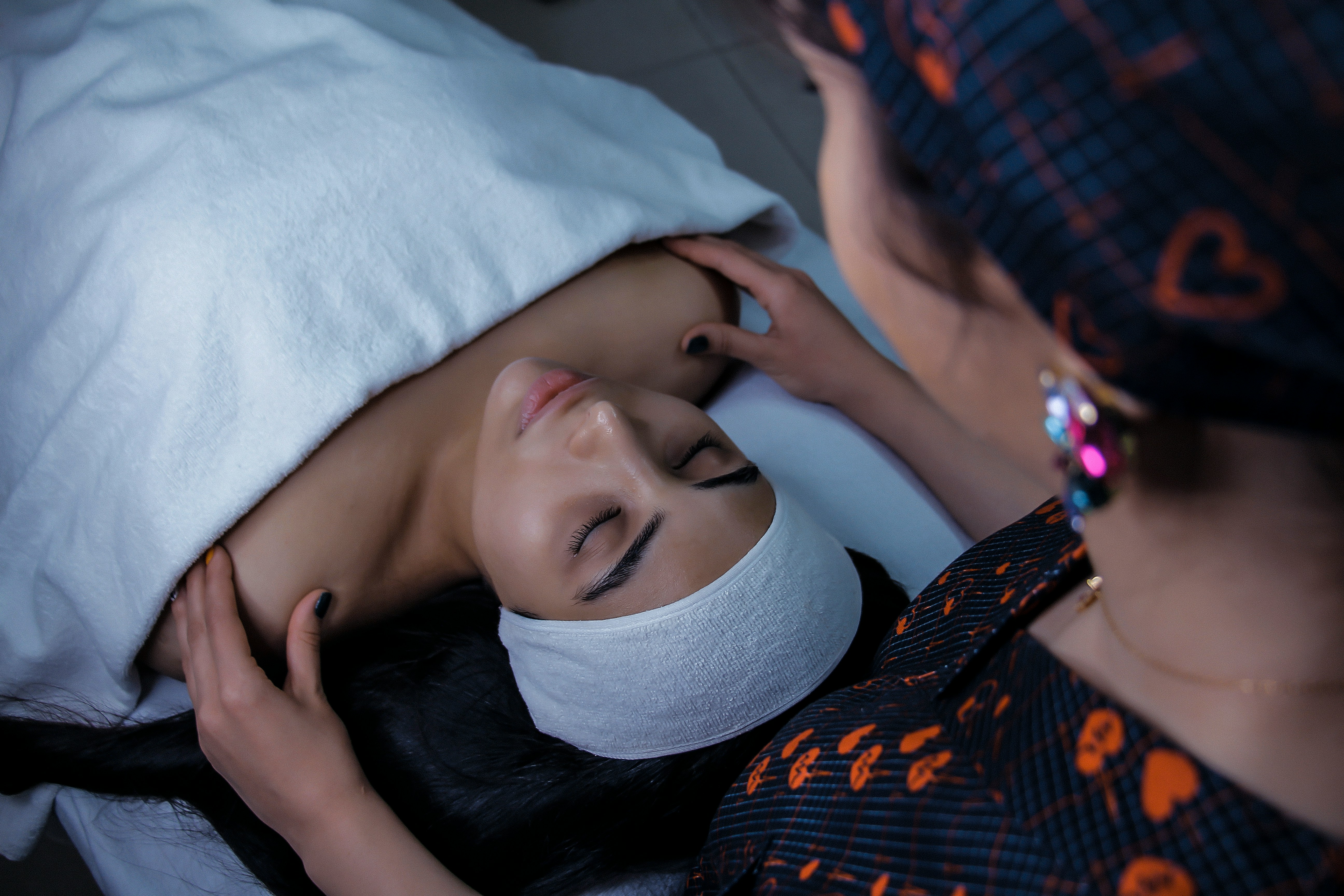 A woman receiving a massage in a spa setting, lying on a massage table with a white towel covering her body and a headband on her head. Another woman is gently placing her hands on the first woman's shoulders.