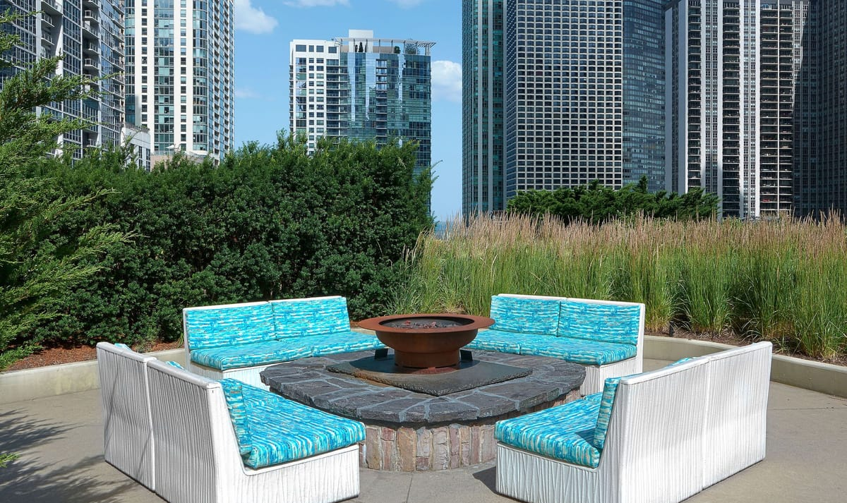 An outdoor lounge area with circular seating around a fire pit, surrounded by tall grass and modern skyscrapers in the background.