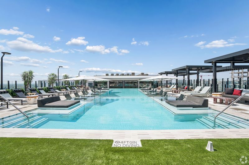 A luxurious rooftop swimming pool area featuring lounge chairs, umbrellas, and greenery, with a clear blue sky above.