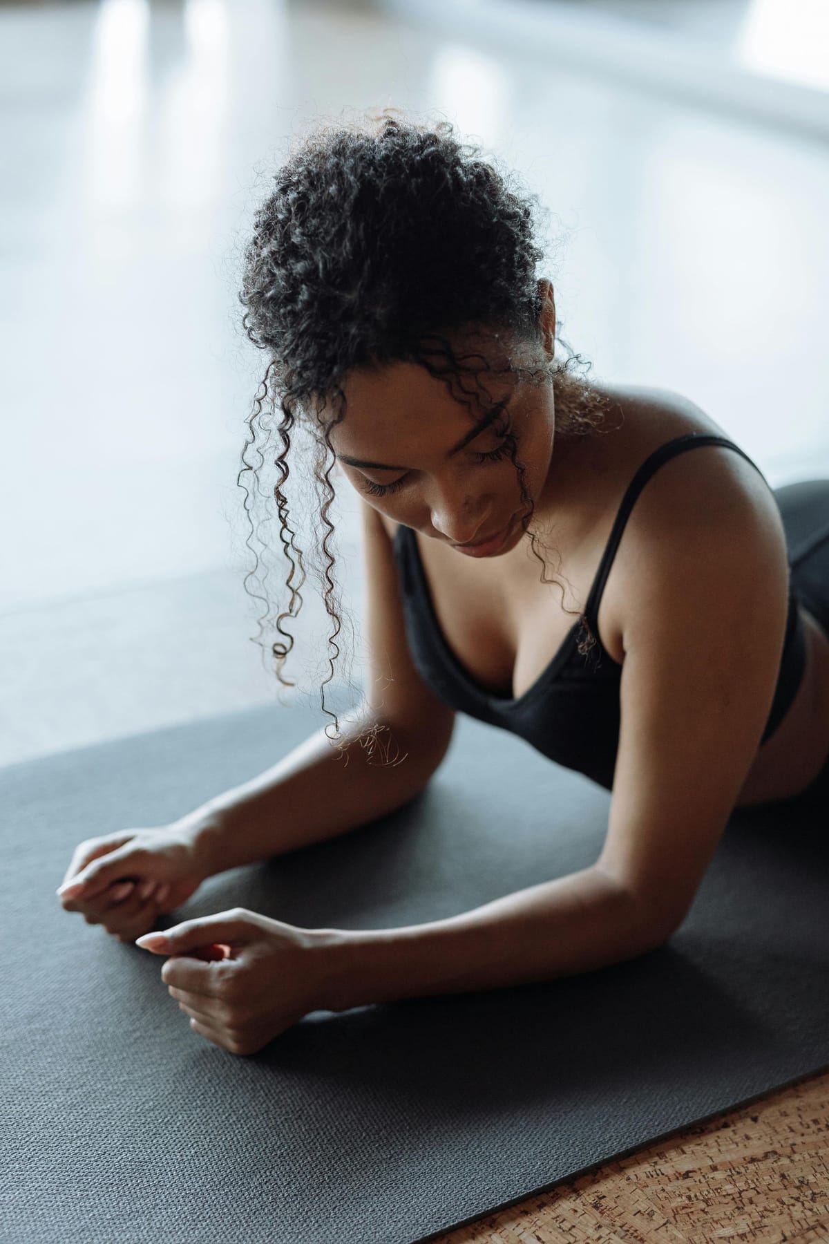 A woman in black sportswear is lying on her stomach on a yoga mat, looking down thoughtfully. The background is softly blurred, highlighting her focused expression.