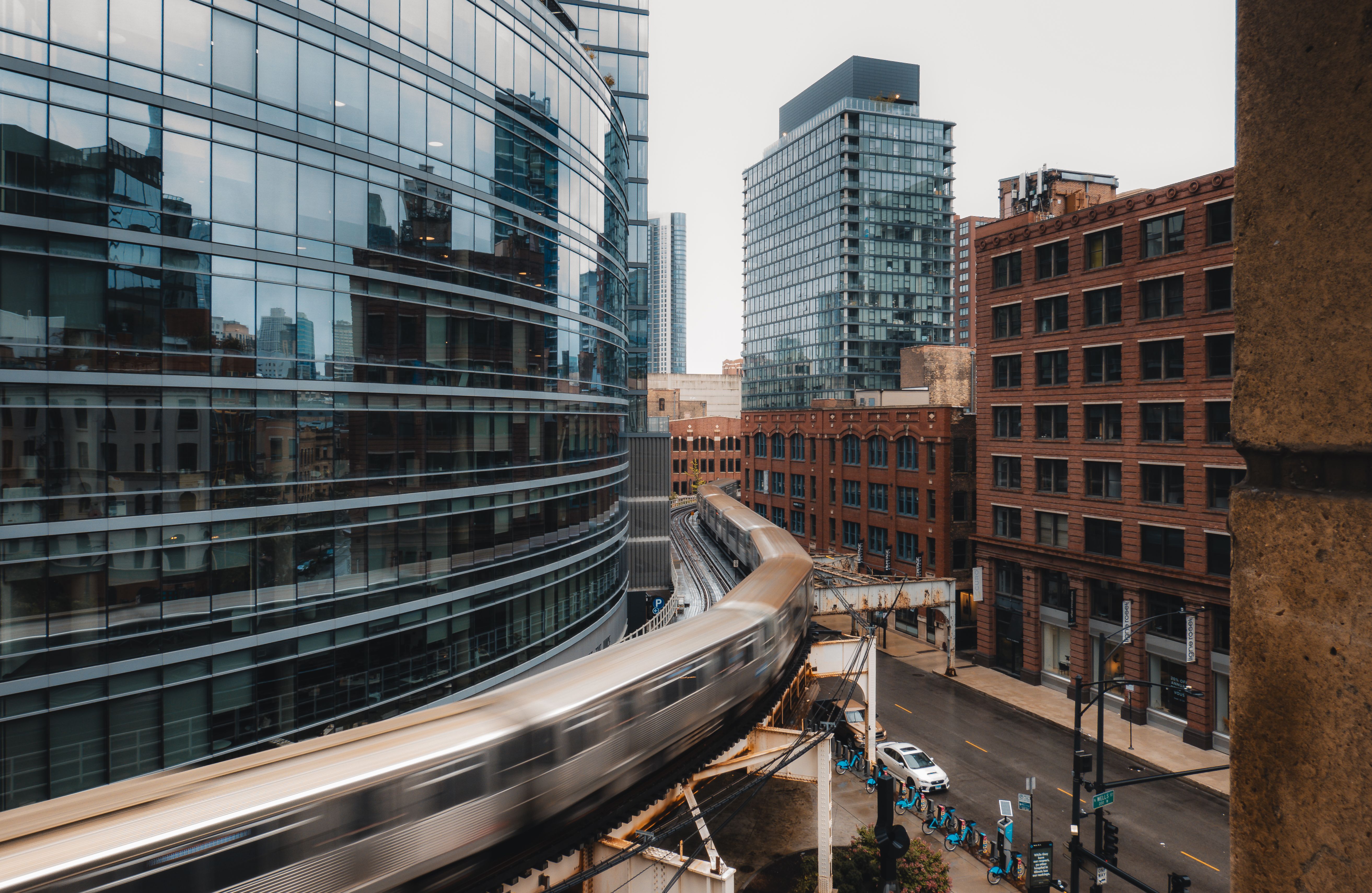 A modern cityscape featuring a curved train track with a train in motion, surrounded by glass buildings and brick structures.