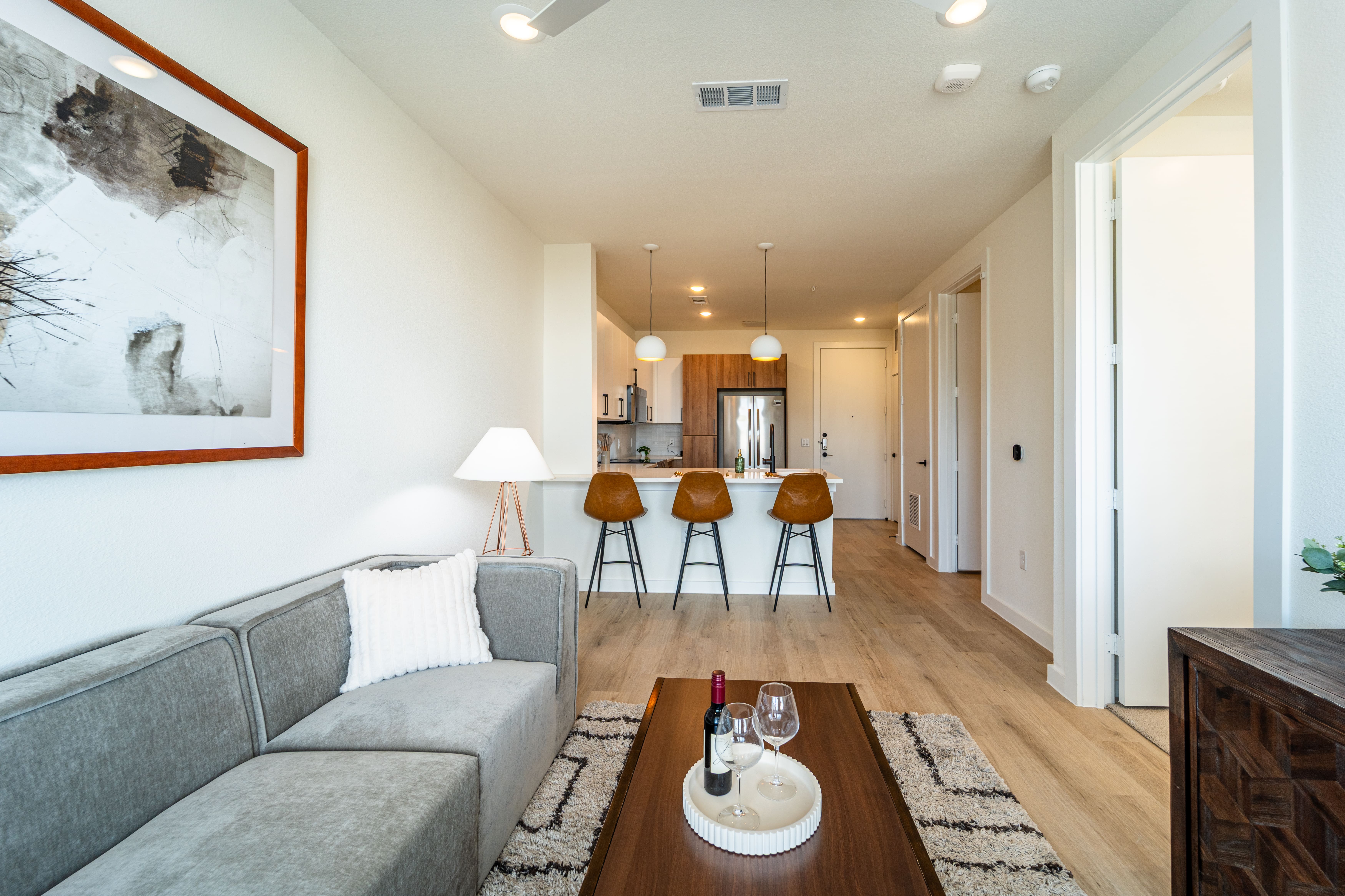 A modern living room featuring a grey sofa, a wooden coffee table with wine glasses, a bottle of wine, and a bowl of snacks. In the background, there are three bar stools and an open kitchen area with pendant lighting. A framed artwork is hanging on the wall.