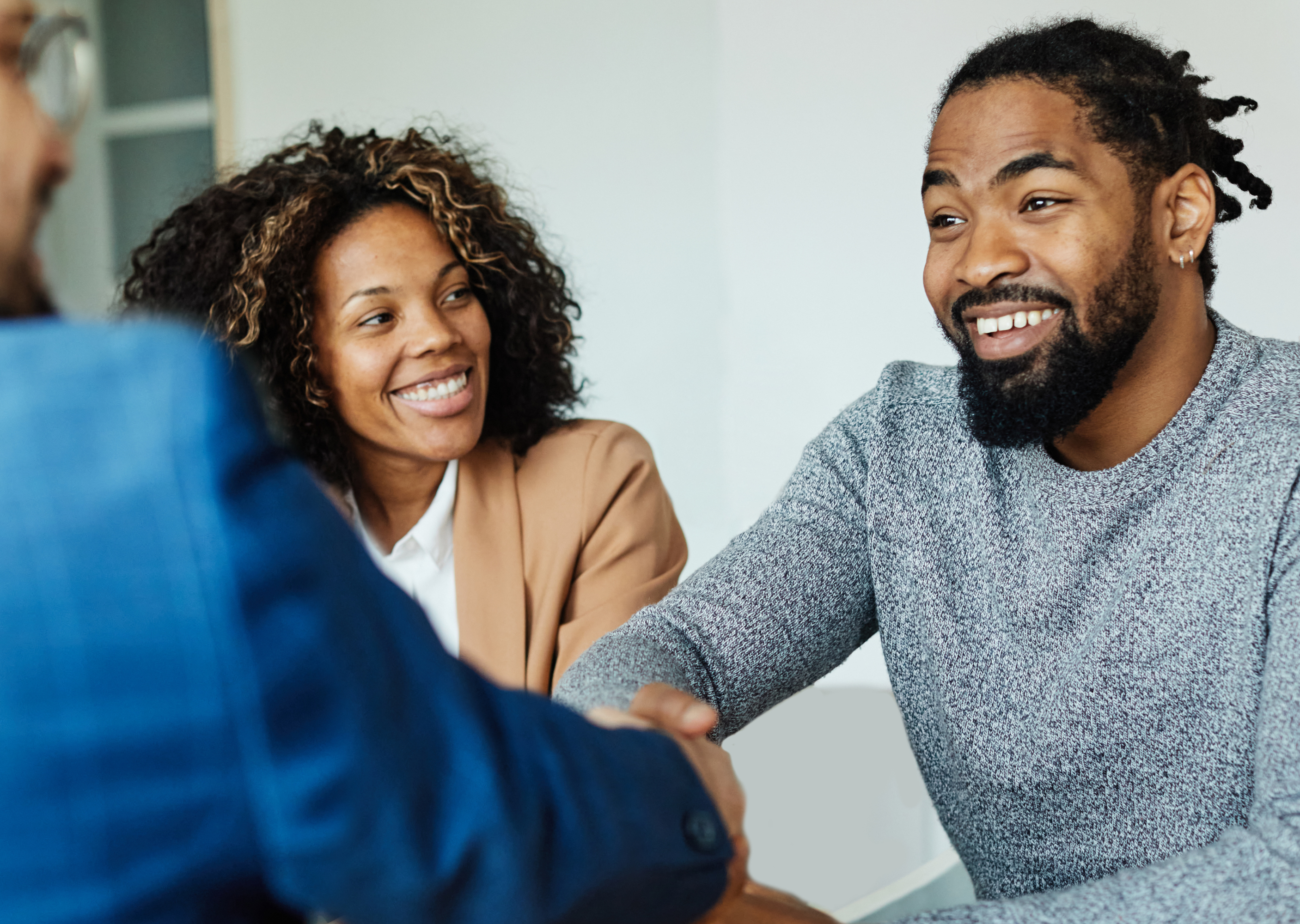 A business meeting involving three individuals. Two people, a woman with curly hair in a beige blazer and a man with a beard in a gray sweater, are smiling as they shake hands with a person in a suit. The setting appears to be a modern office.