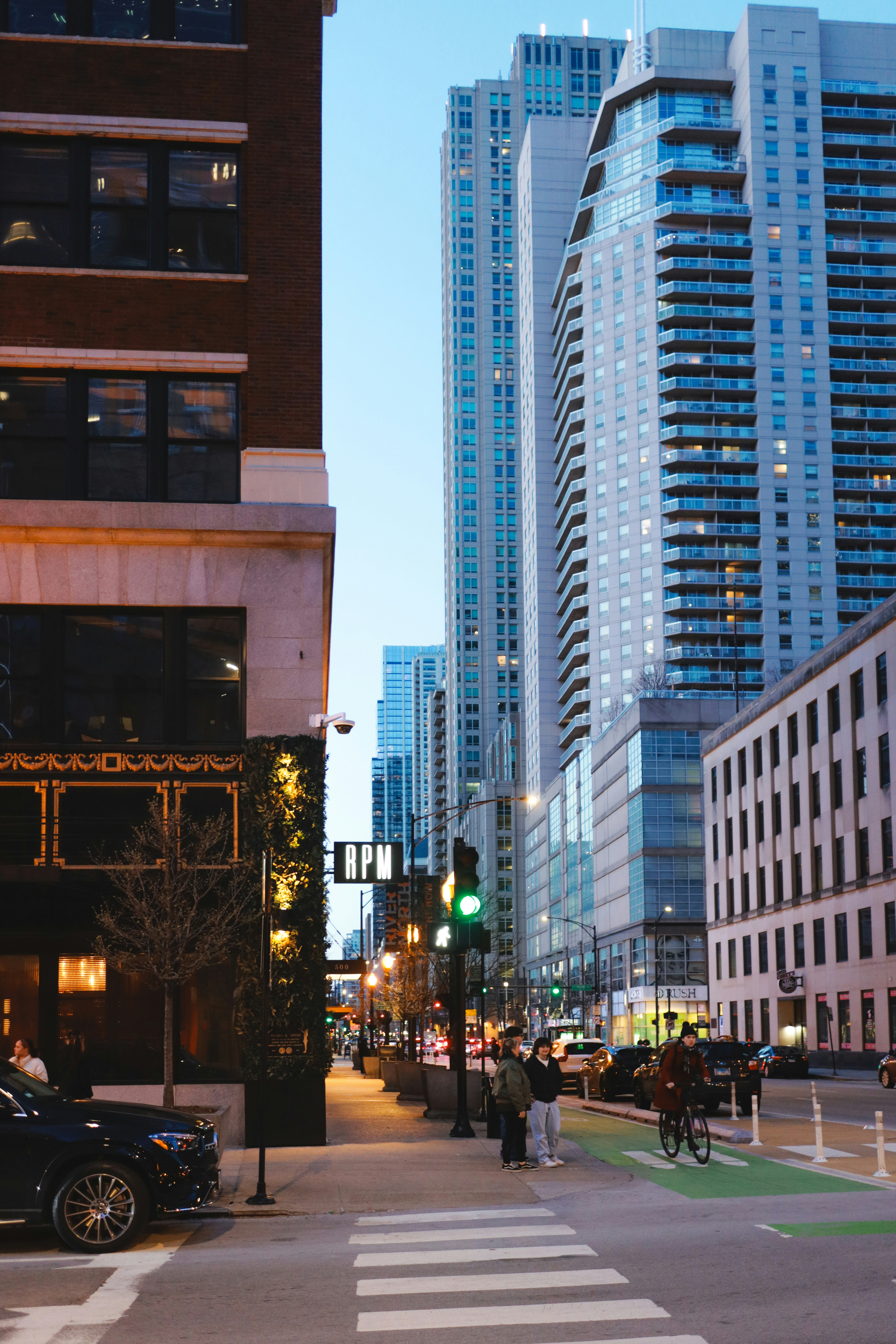 A city street view during evening time featuring tall buildings, a street with green bike lanes, and people walking.