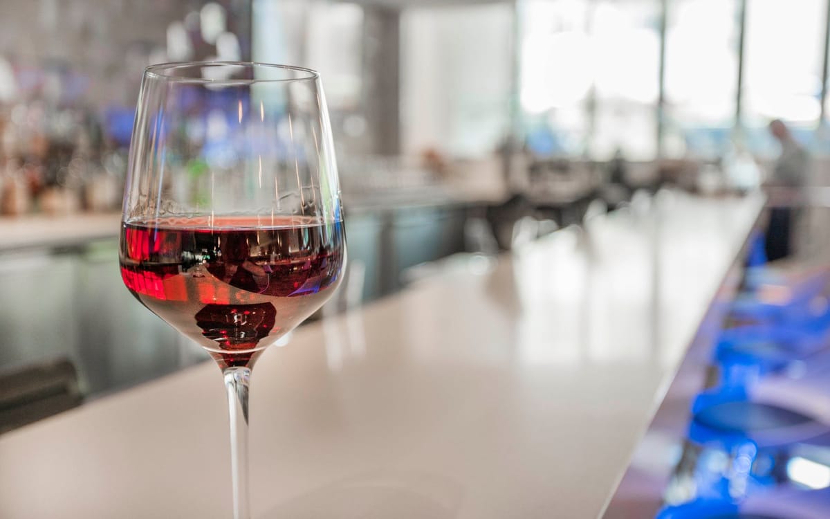 Dining, Cafes, and Culinary Delights | A close-up of a glass of red wine placed on a sleek bar counter with blurred background details of a modern bar.