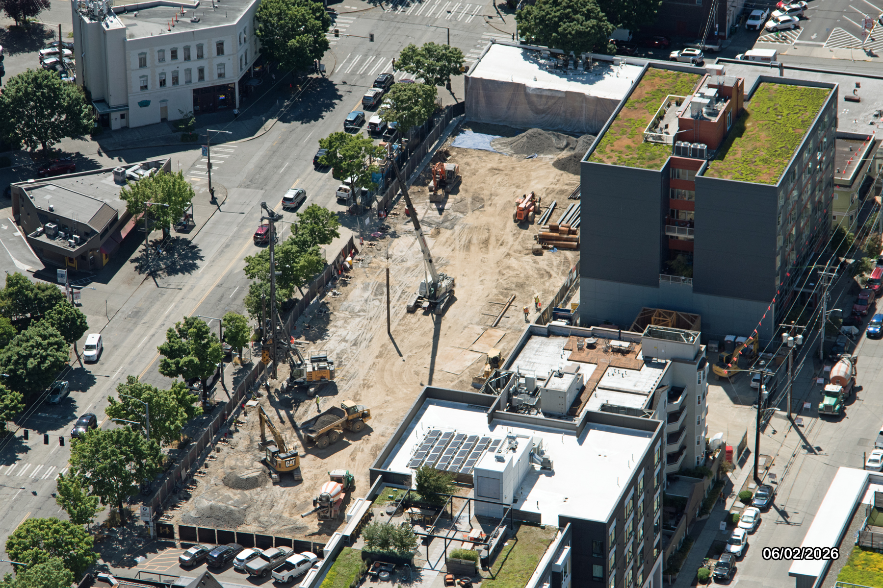 Aerial view of a construction site surrounded by roads and buildings. Heavy machinery is present on a cleared area with construction materials.