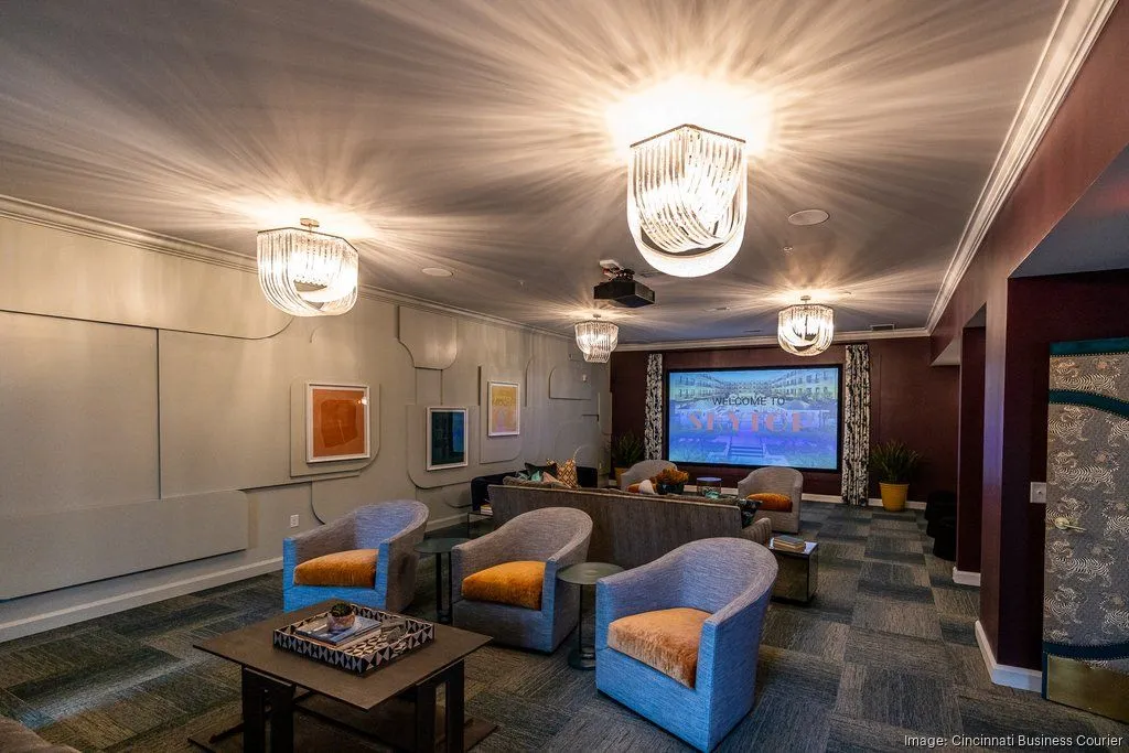 A modern lounge area featuring comfortable blue chairs with orange cushions, a coffee table, and a large screen at the far end. The space is well-lit with stylish chandeliers and has a contemporary aesthetic.