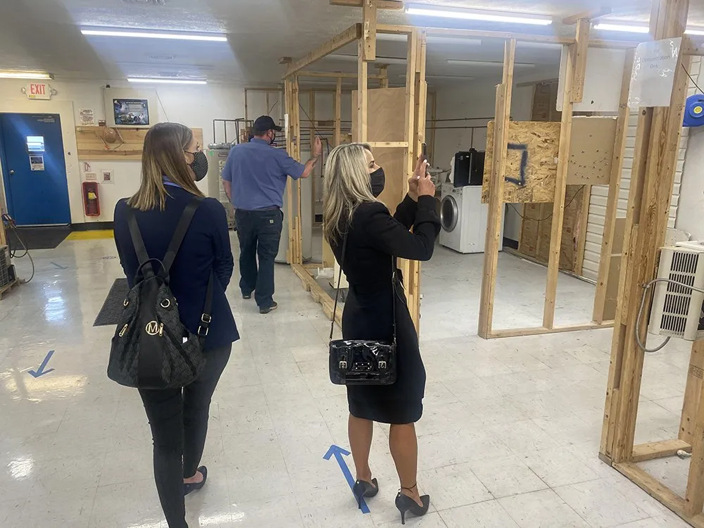 Two women wearing masks are touring a partially constructed apartment space with wooden frames; one is taking a photo while the other observes.