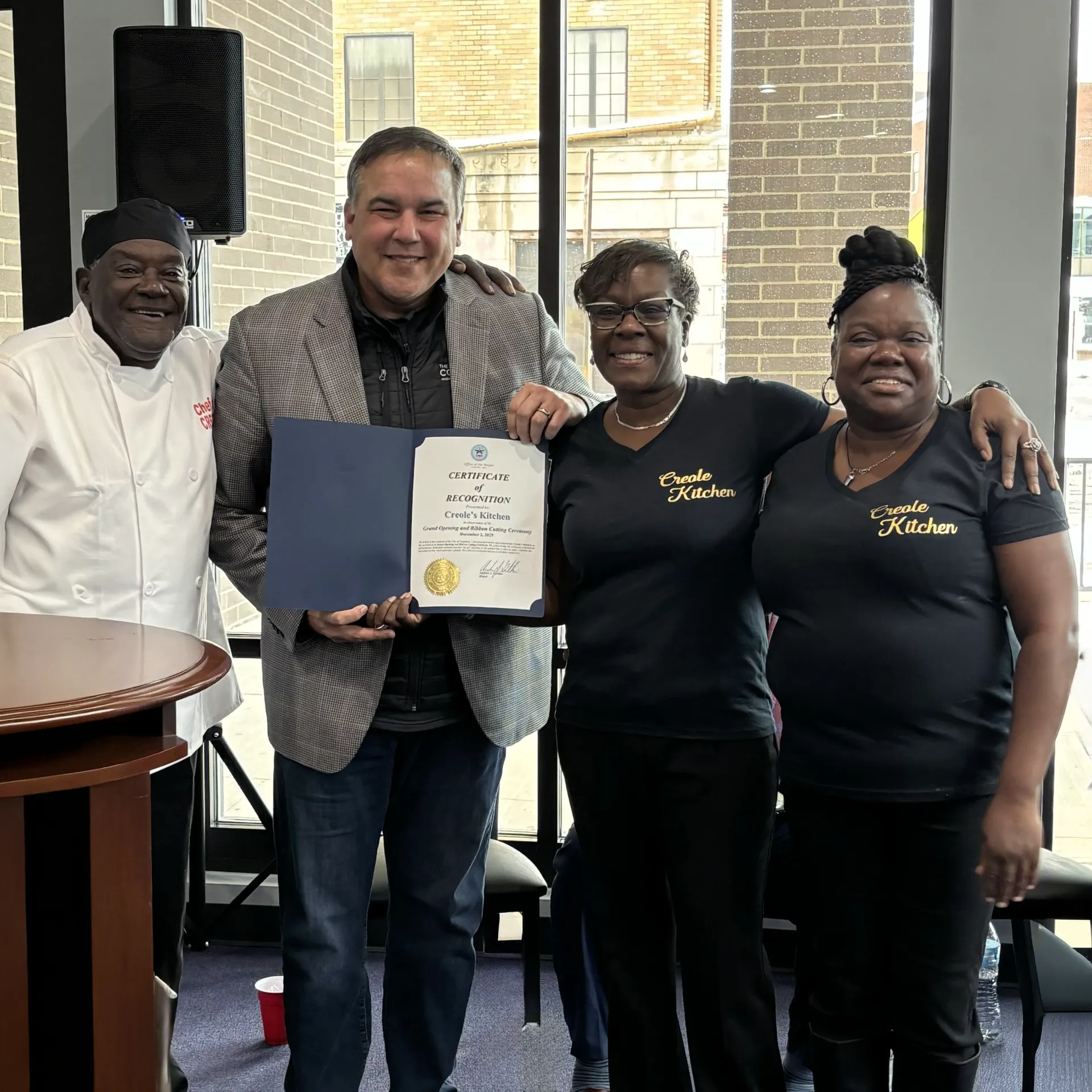 Group of four people smiling together at an event holding a certificate, with one person in a chef's uniform and others in matching shirts.