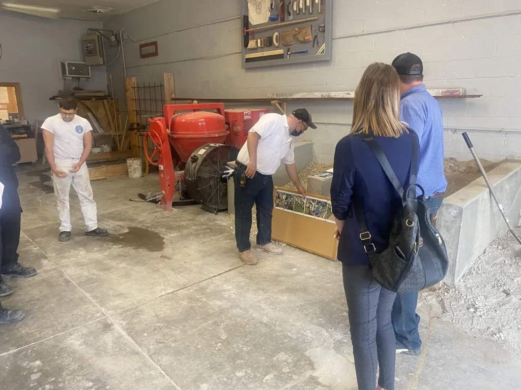 A construction training session taking place in a garage or workshop. Two people observe a demonstration involving a construction mixer and a cardboard box on the floor. The environment includes various tools and equipment.