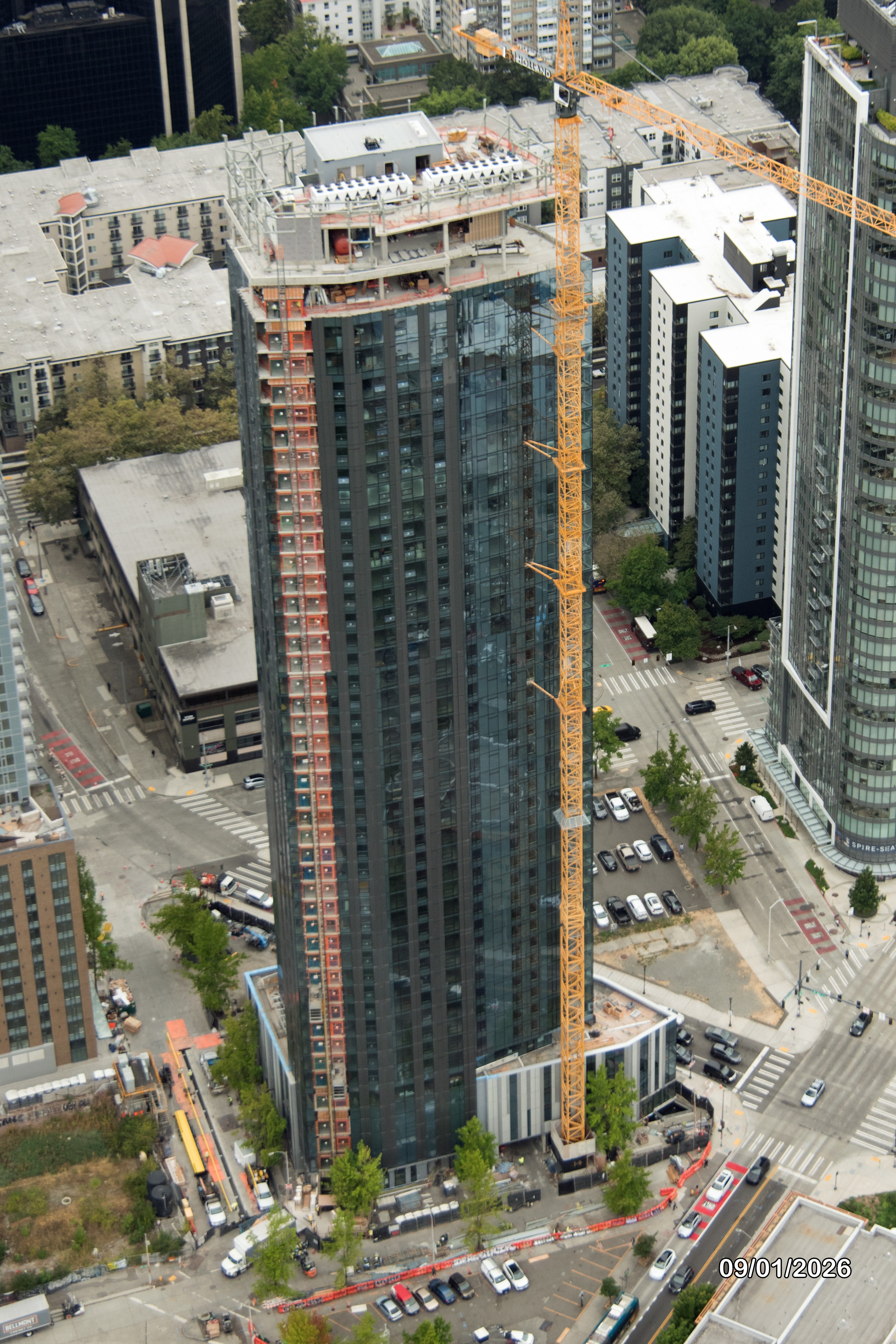 An aerial view of a high-rise residential building under construction, featuring scaffolding and a crane, with surrounding urban development and streets visible.