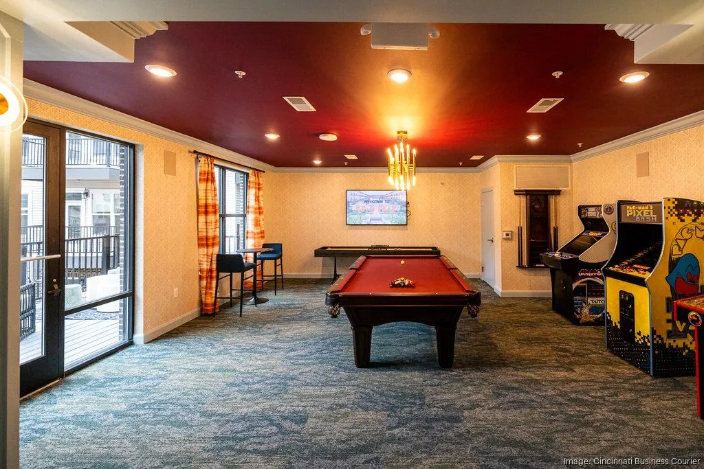 A community game room featuring a pool table, arcade machines, and seating areas, with large windows and colorful decor.