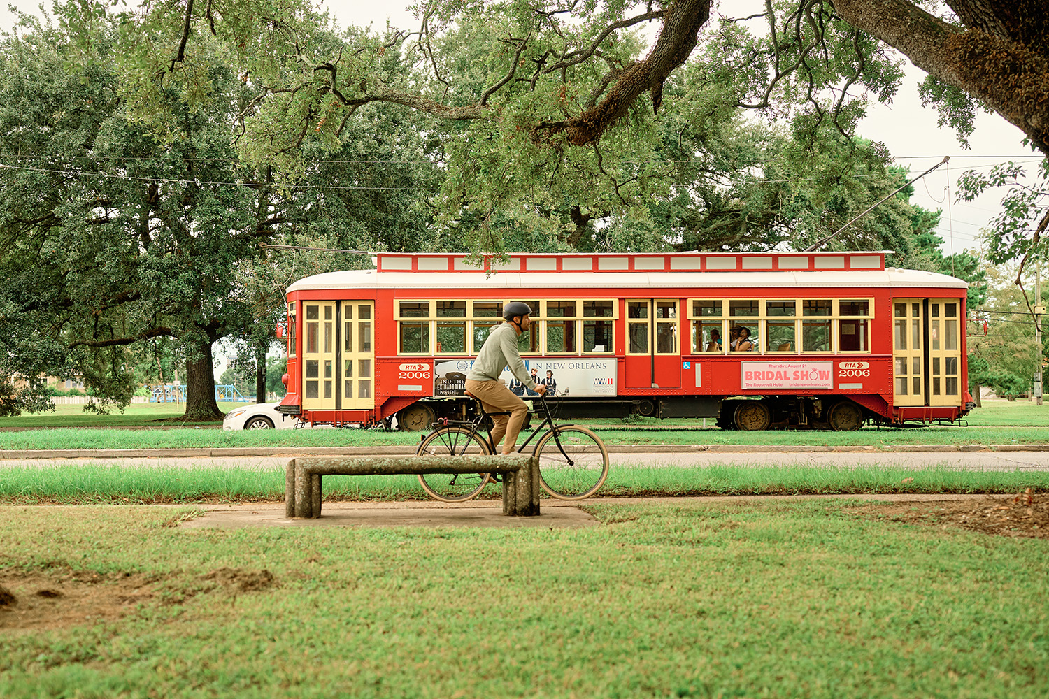 A person riding a bicycle past a red tram on a green grassy area, with trees in the background.