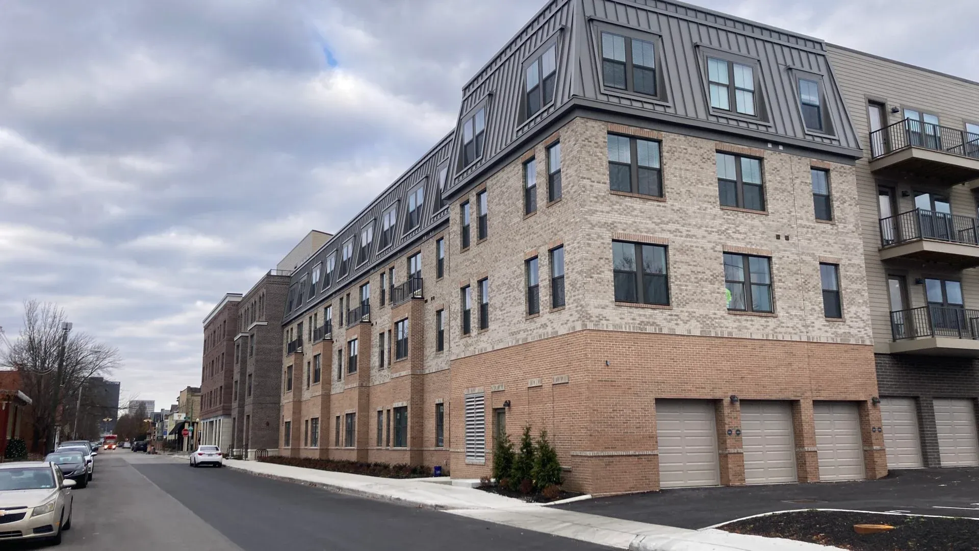A modern multi-story apartment building with a brick facade and large windows, situated on a street with cars parked along the side.