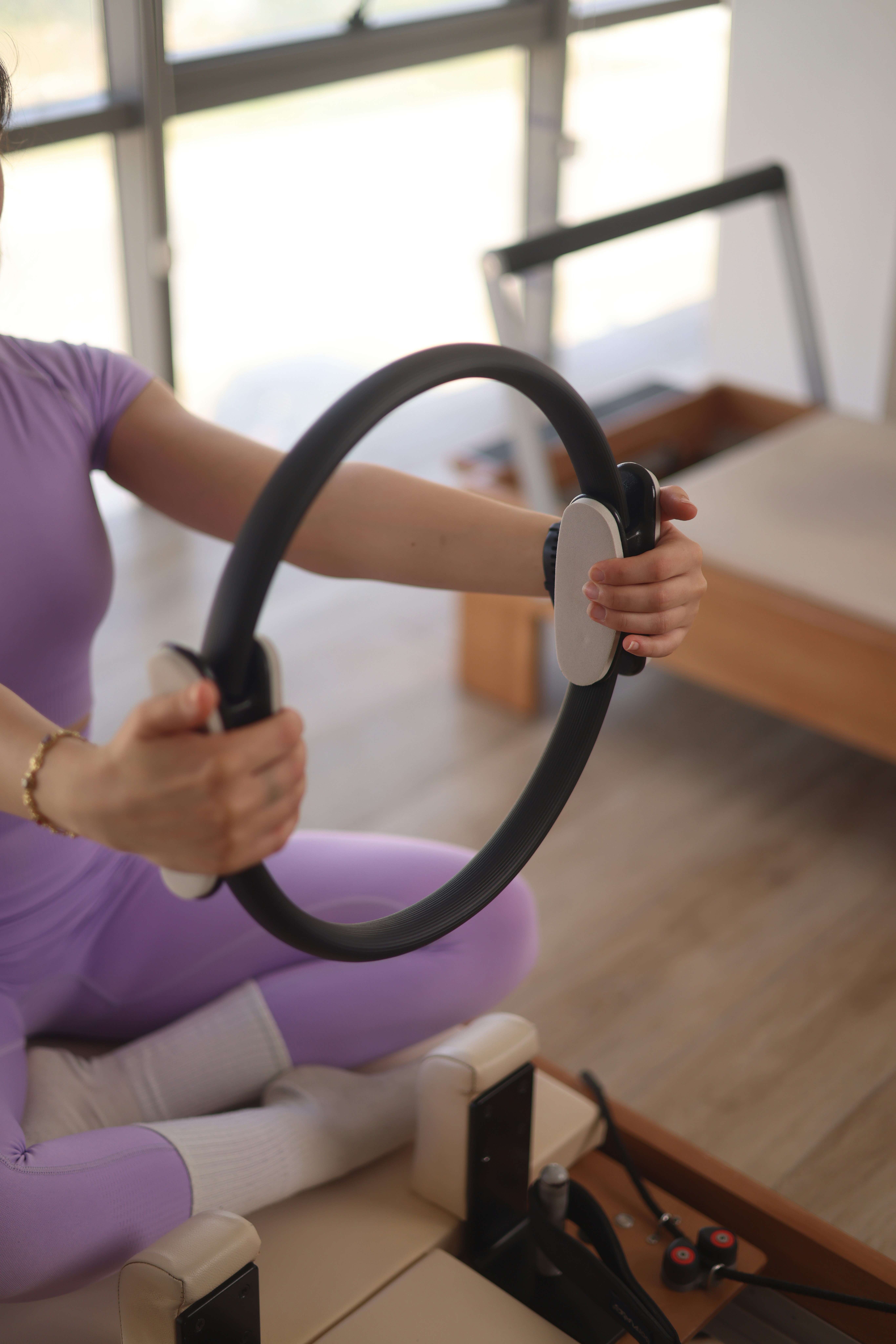 A person in a purple outfit performing an exercise with a Pilates ring, sitting on a reformer in a bright room.
