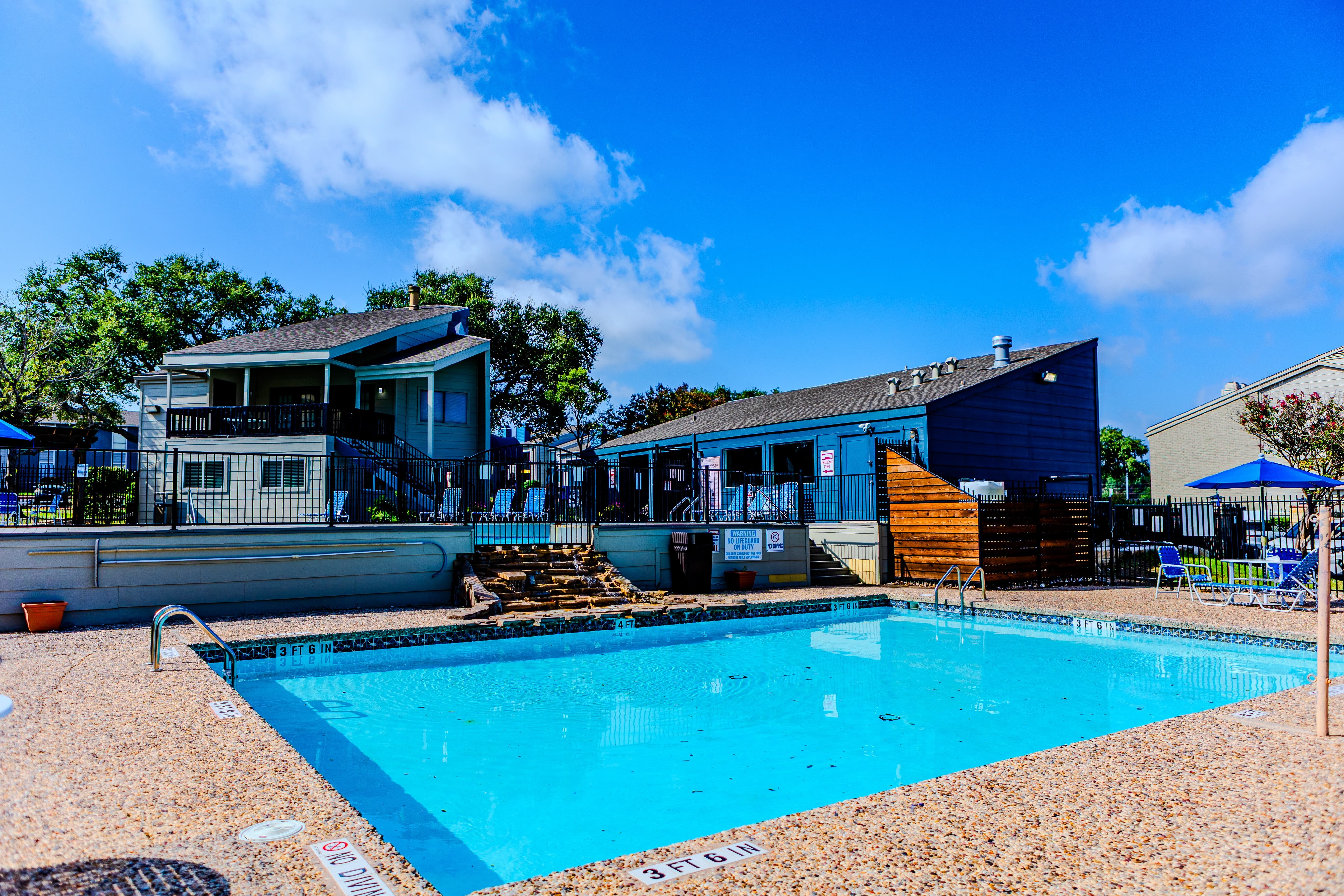 A sunny outdoor pool area with a clear blue sky, surrounded by two multi-story residential buildings and lounging chairs.