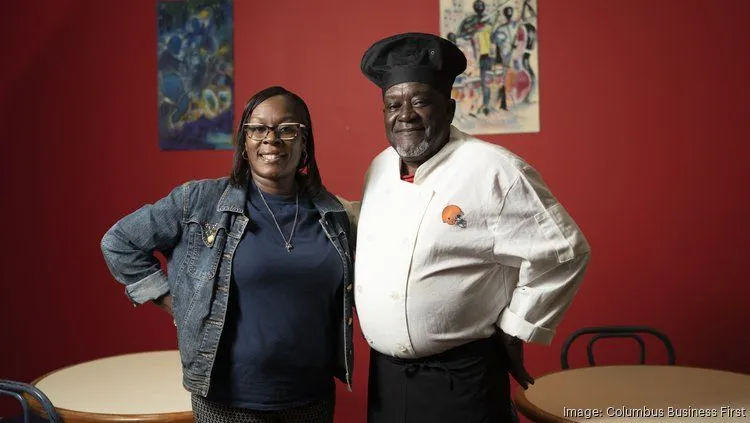 A woman and a man wearing a chef's uniform stand smiling together in a restaurant with a red background and colorful artwork on the walls.