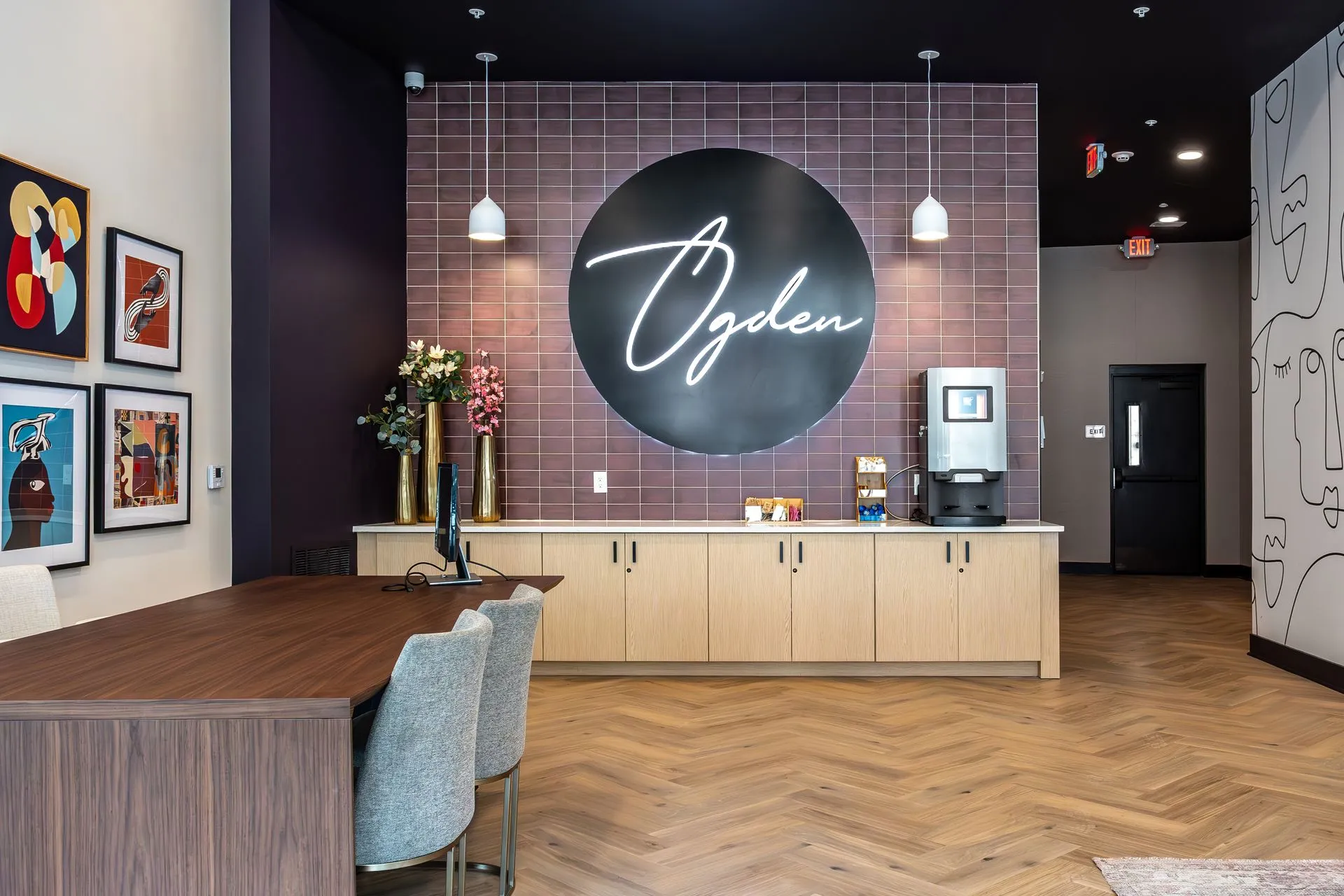 A modern lobby area featuring a welcoming design, with a large round wall sign that reads 'Ogden' and a beverage station. The space has wooden flooring and contemporary artwork on the walls.