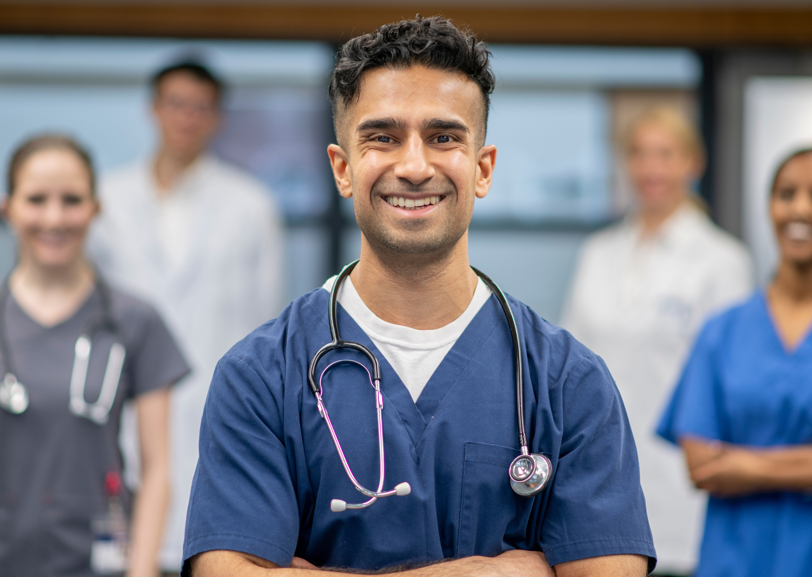 A healthcare professional in scrubs smiles confidently in the foreground, surrounded by other medical staff in the background. The setting appears to be a medical facility or hospital.